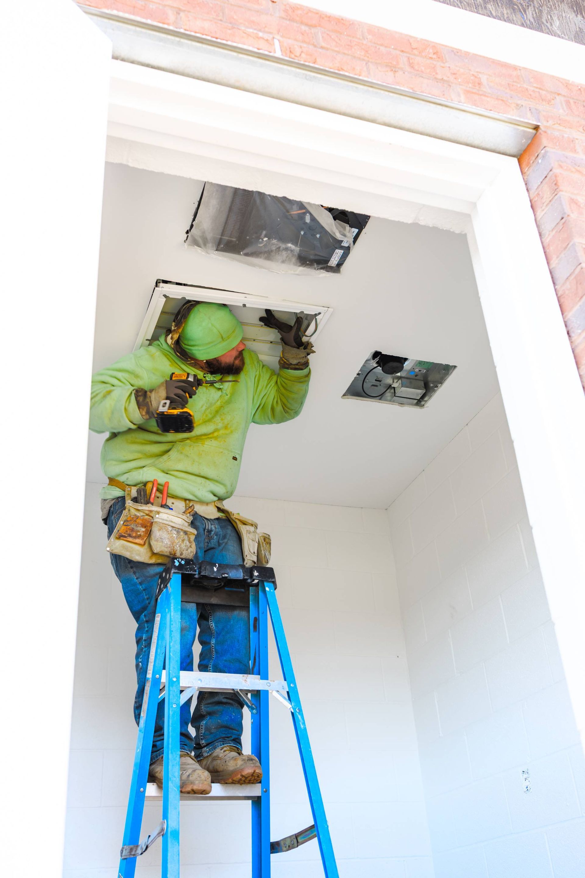 Construction worker on a ladder, installing ceiling fixtures. Green hoodie, blue ladder, brick building.