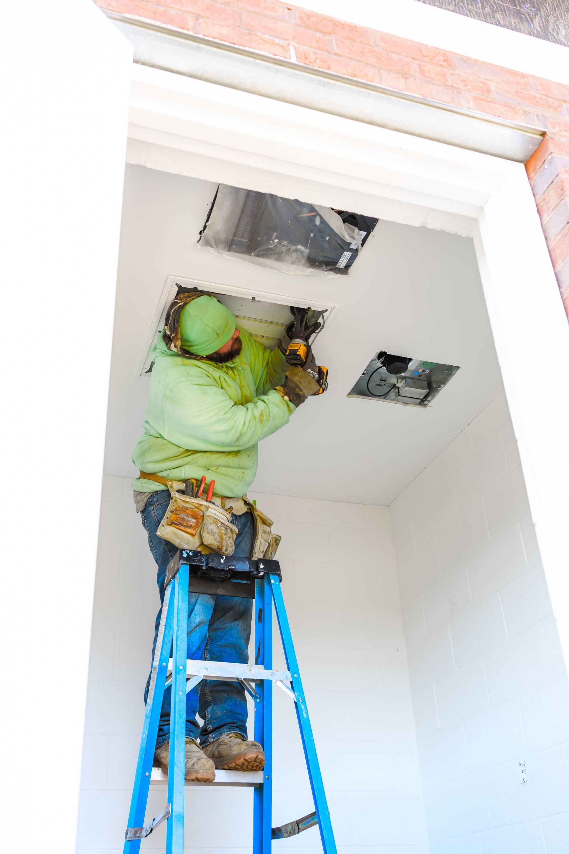 Construction worker on a blue ladder installing something into a ceiling opening. White walls and a brick exterior.
