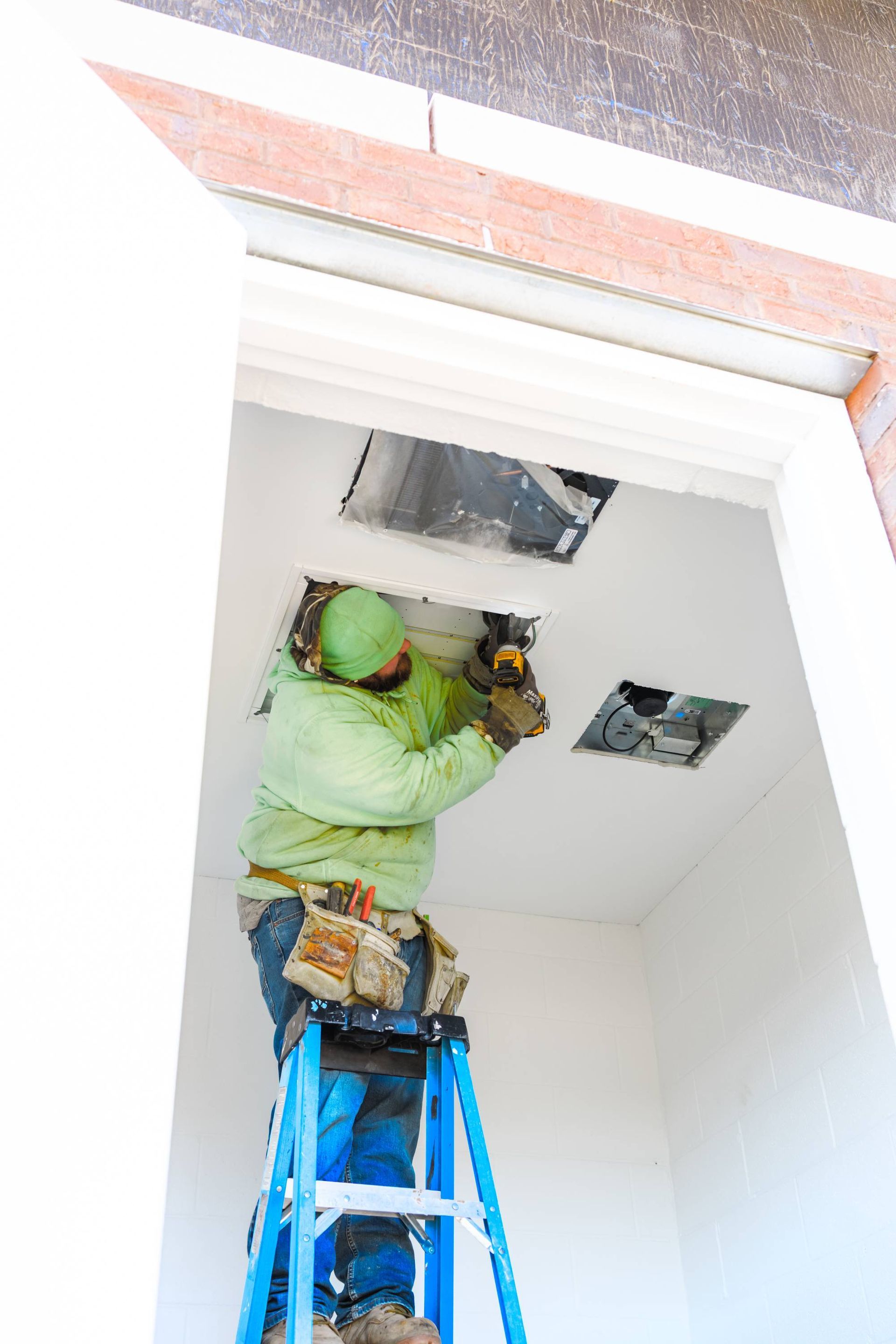 Construction worker on a ladder, installing something into a ceiling. Green jacket, blue ladder, white walls.