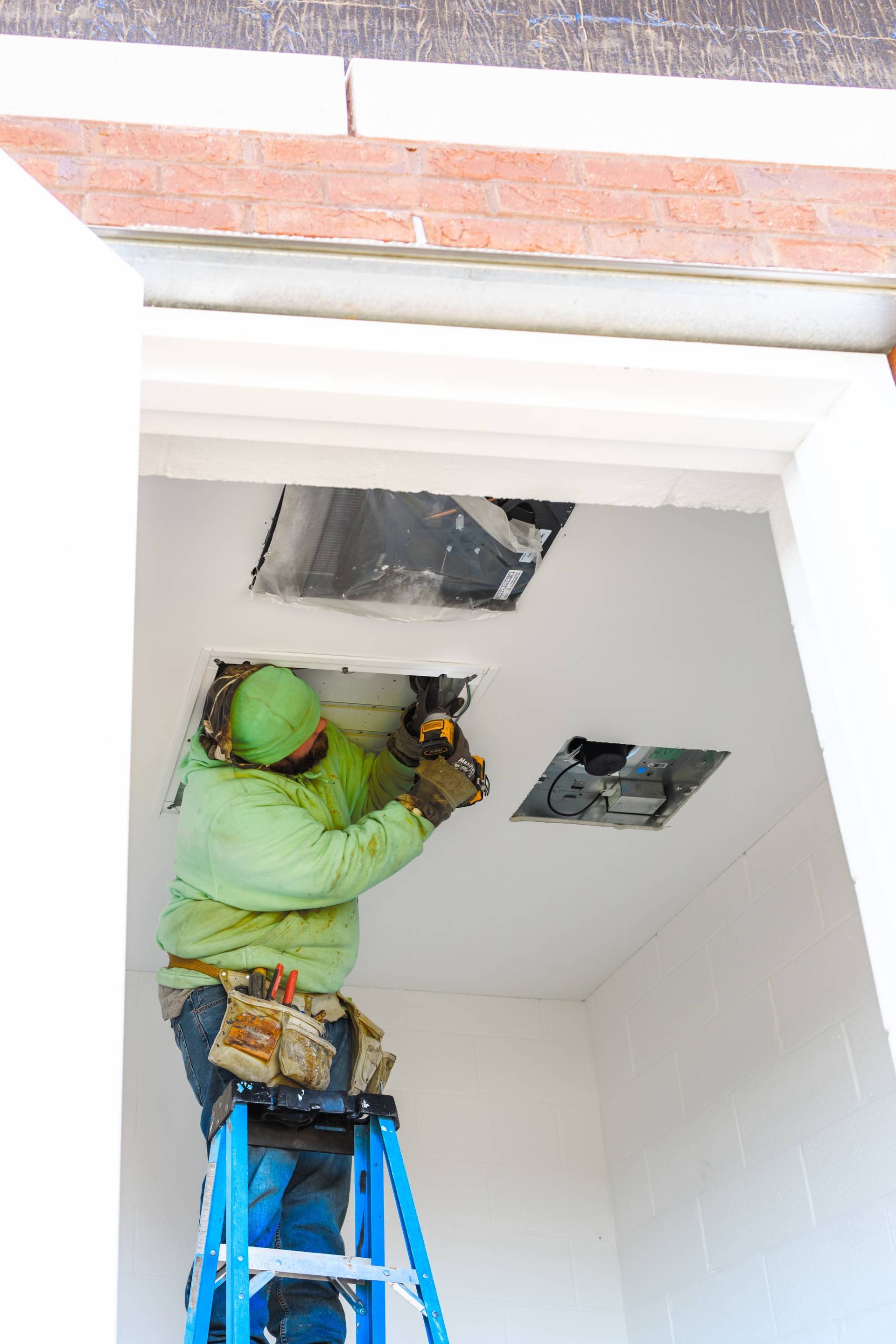 A worker on a ladder installs something in a ceiling. White walls, brick above, and a green jacket.