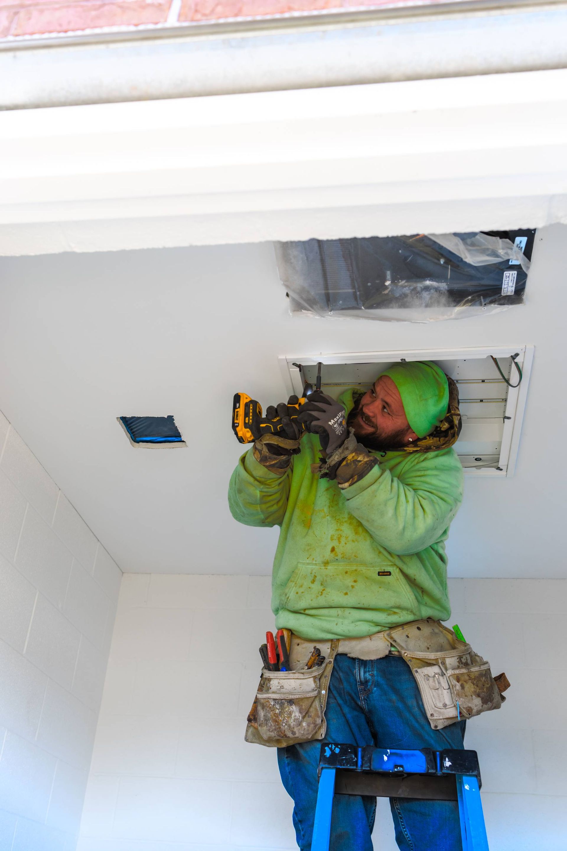 Person on a ladder installing something in a ceiling. He wears a green hat and jacket, and a tool belt.