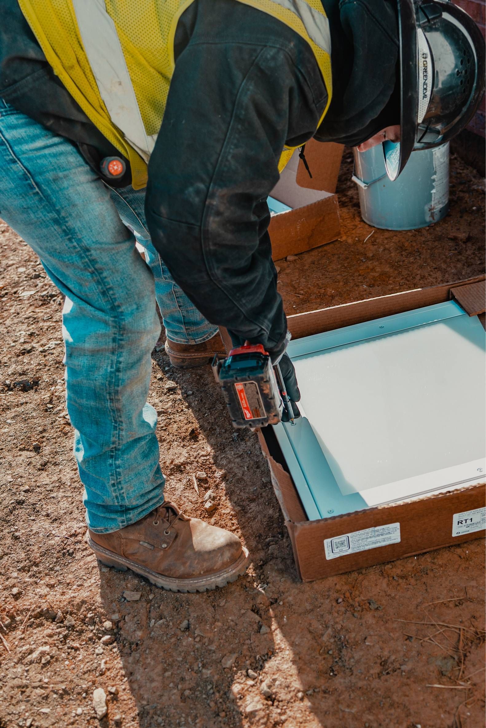 Construction worker using a power tool to assemble a white panel in a cardboard box on a job site.
