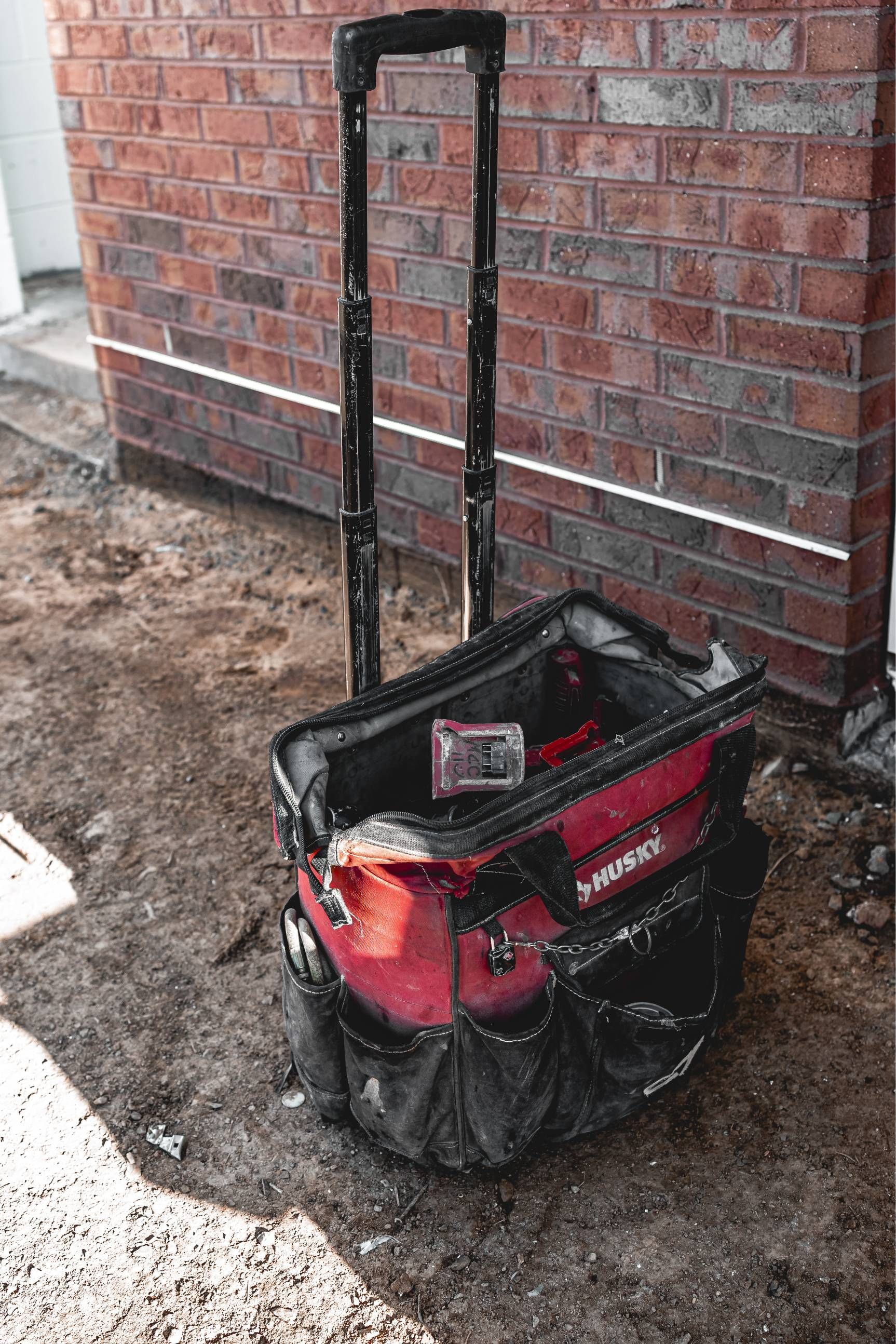 Red rolling tool bag on a dirty surface in front of a brick wall. Black handle is extended.