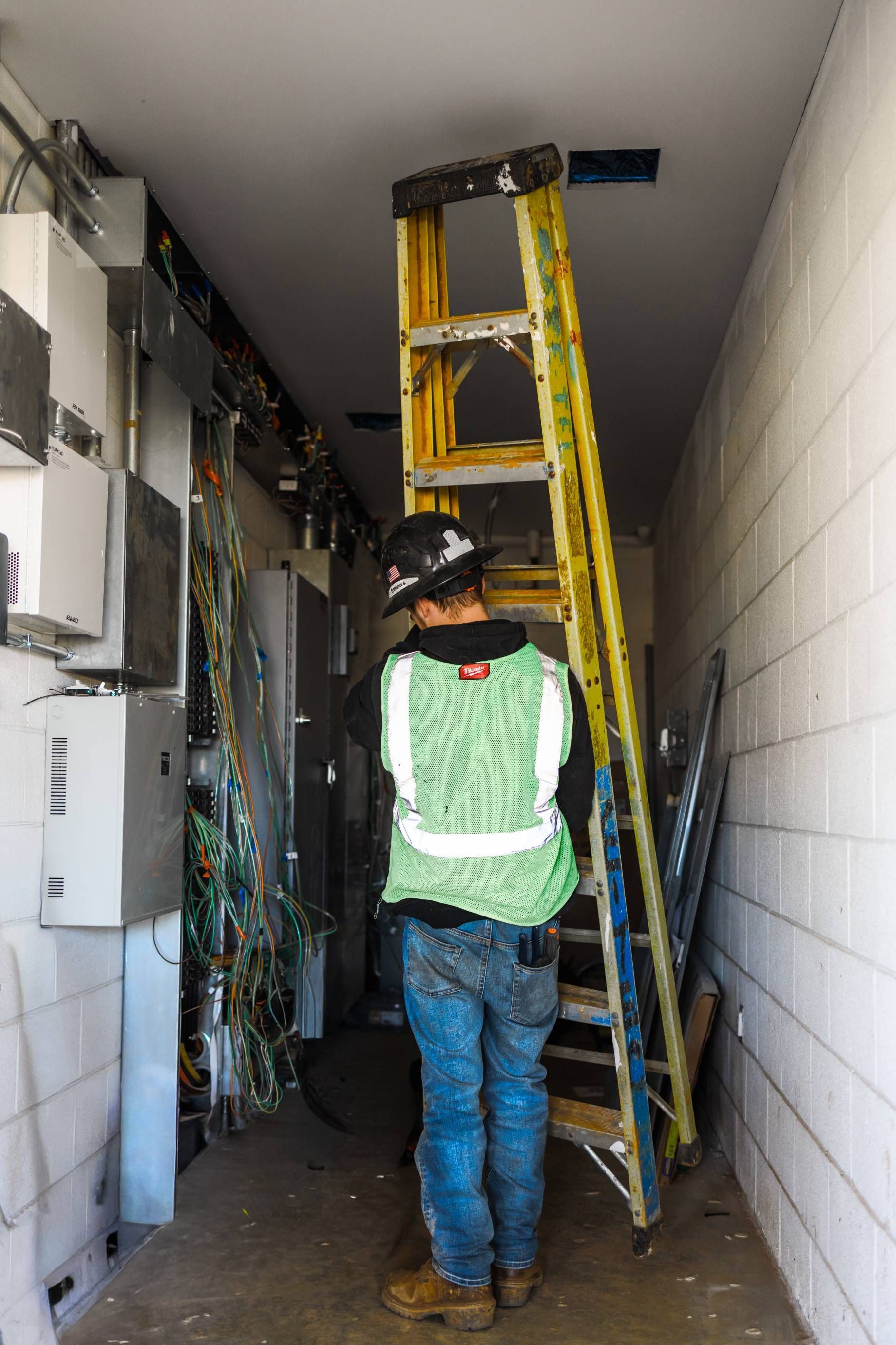 Worker in safety vest stands near ladder in narrow room with wires and electrical boxes.