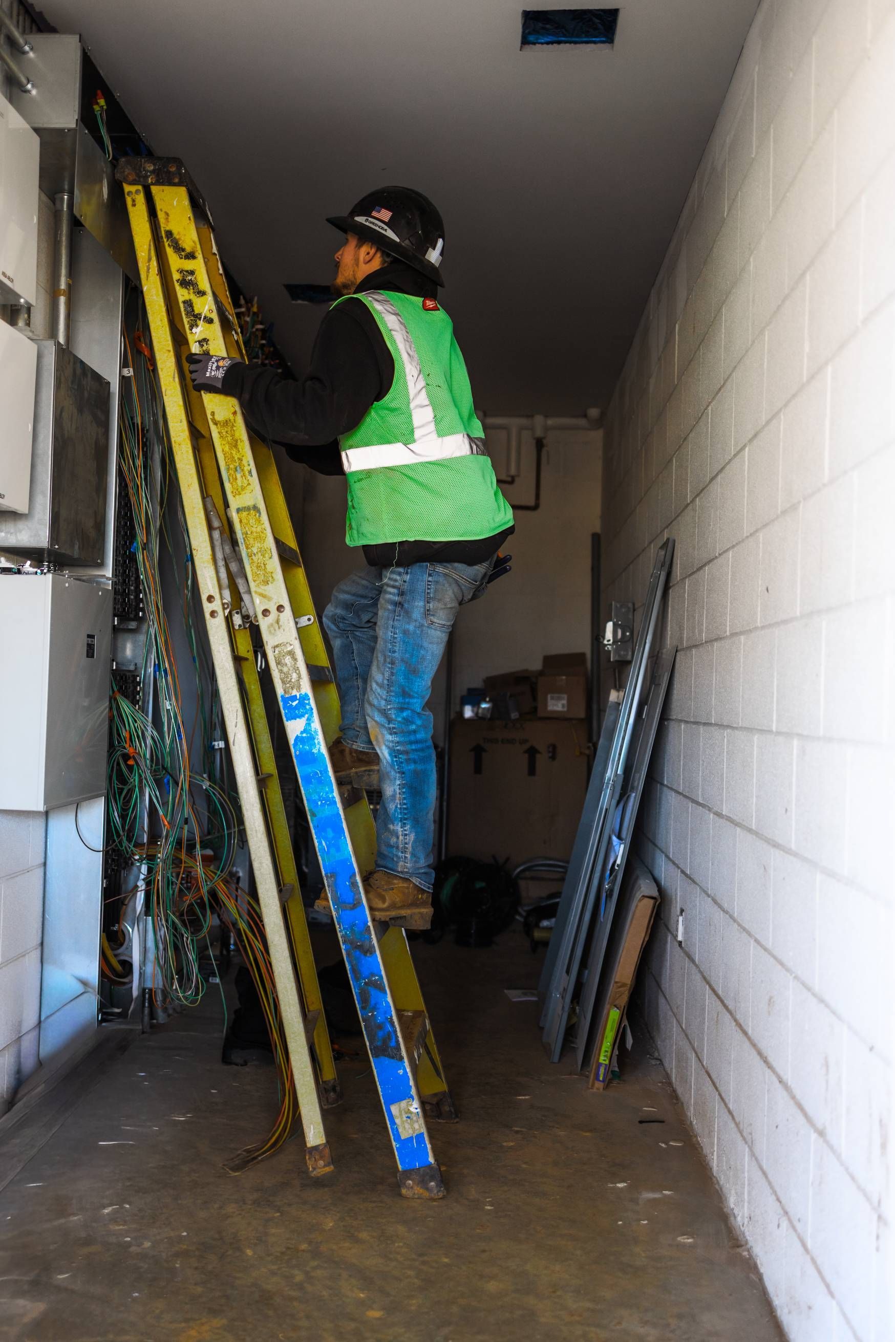 Person wearing a reflective vest climbs a ladder in a narrow space with equipment and cables on the wall.
