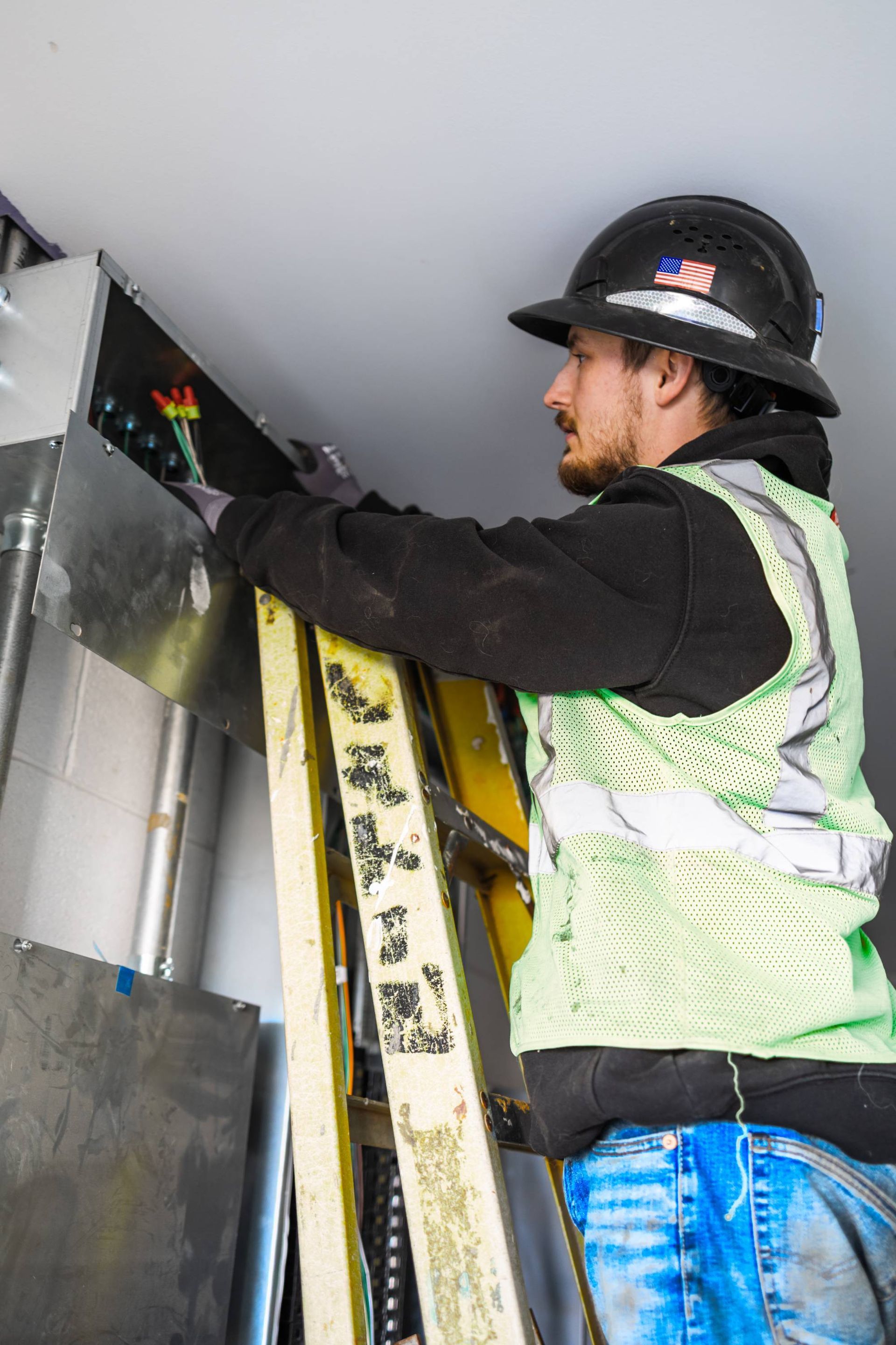 Construction worker on a ladder installing ductwork; wears a hard hat and safety vest, working indoors.