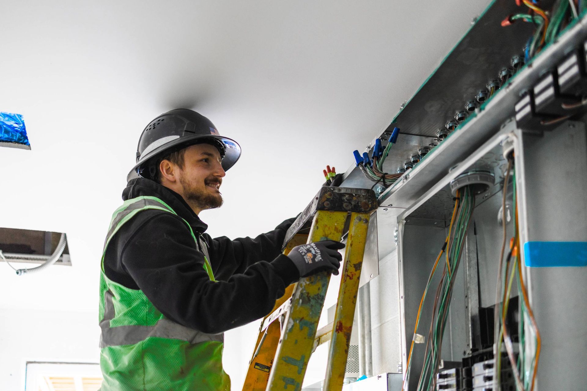 Electrician on ladder working on wires inside a panel. He is wearing a hard hat and safety vest.