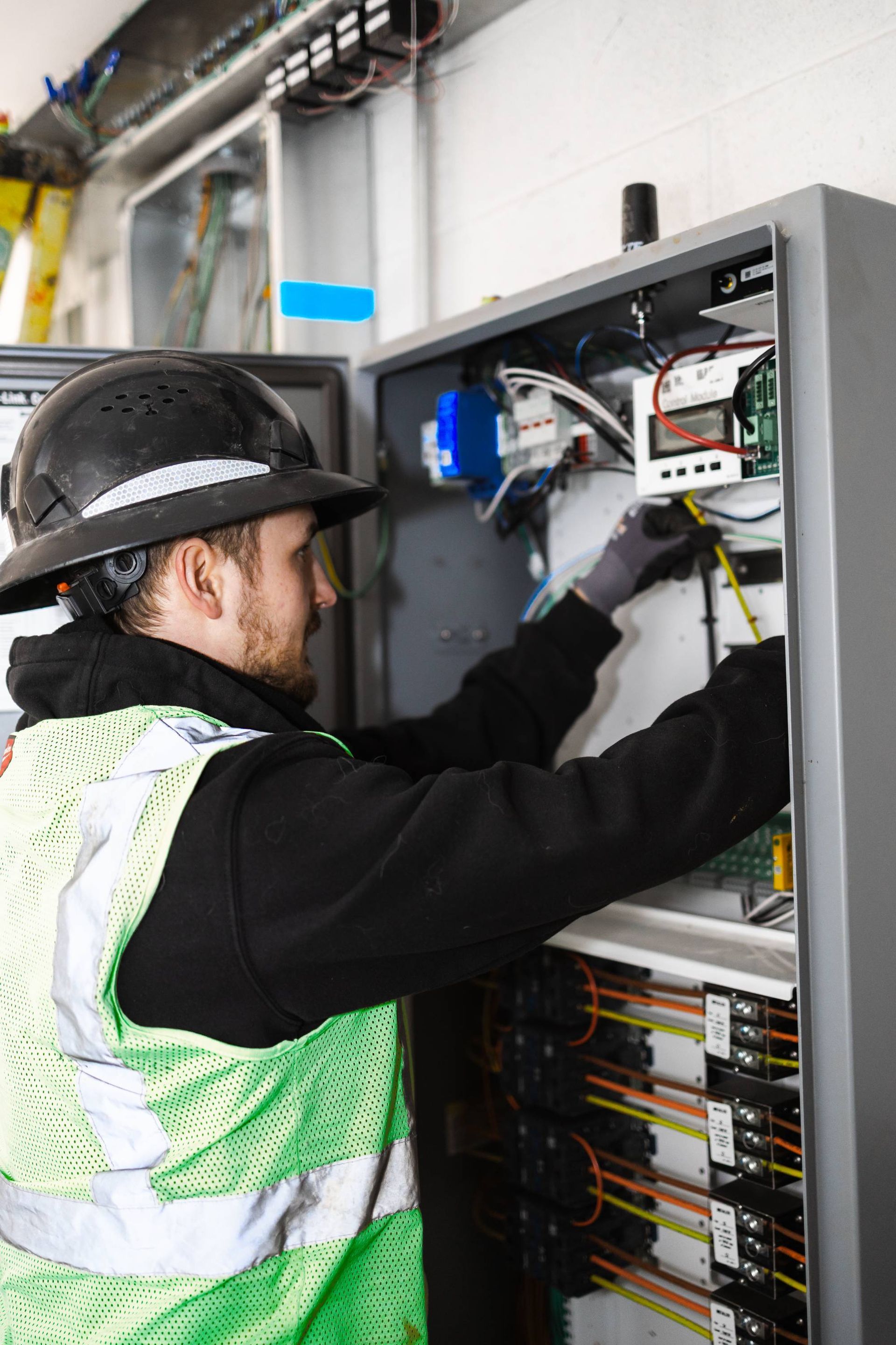 Electrician working on electrical panel, wearing a hard hat and safety vest, indoors.