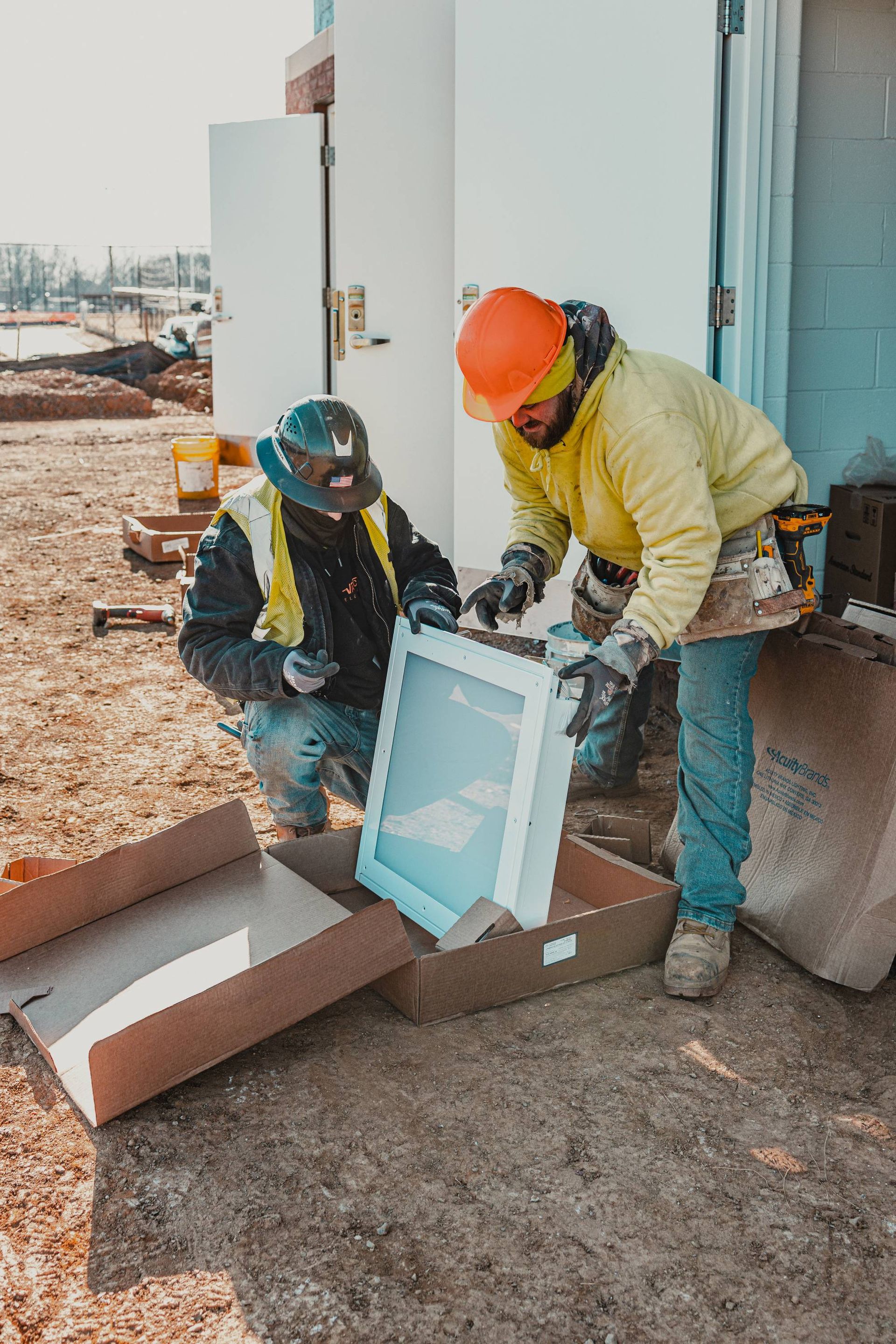 Two construction workers unpacking a light fixture on a job site. One kneels, the other stands.