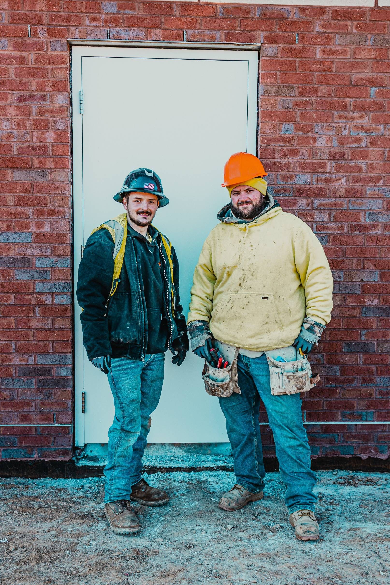 Two construction workers stand by a white door in front of a brick building. One wears a hard hat, the other an orange one.