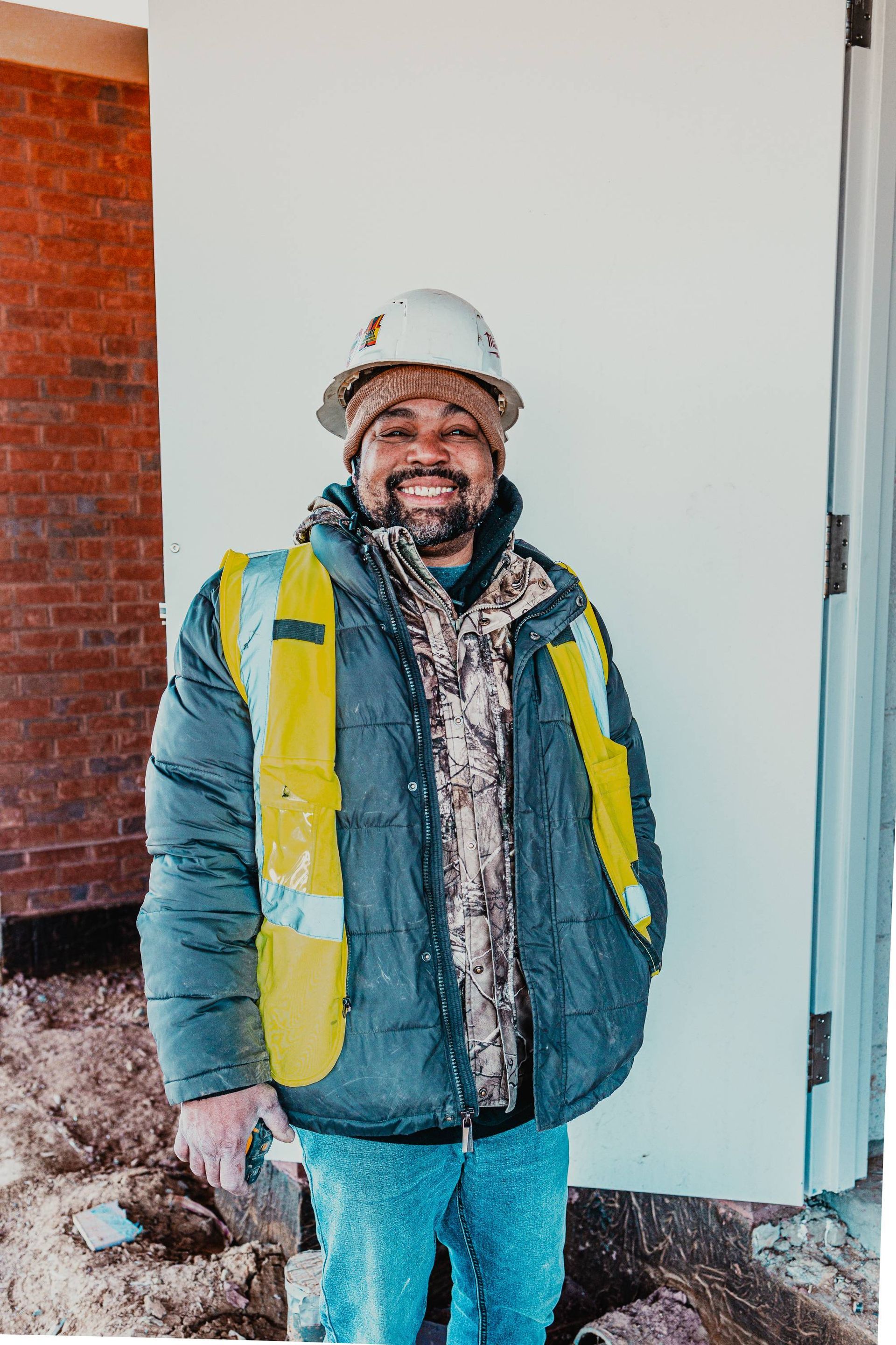 Construction worker in hard hat, safety vest, and winter coat smiling in front of building materials.