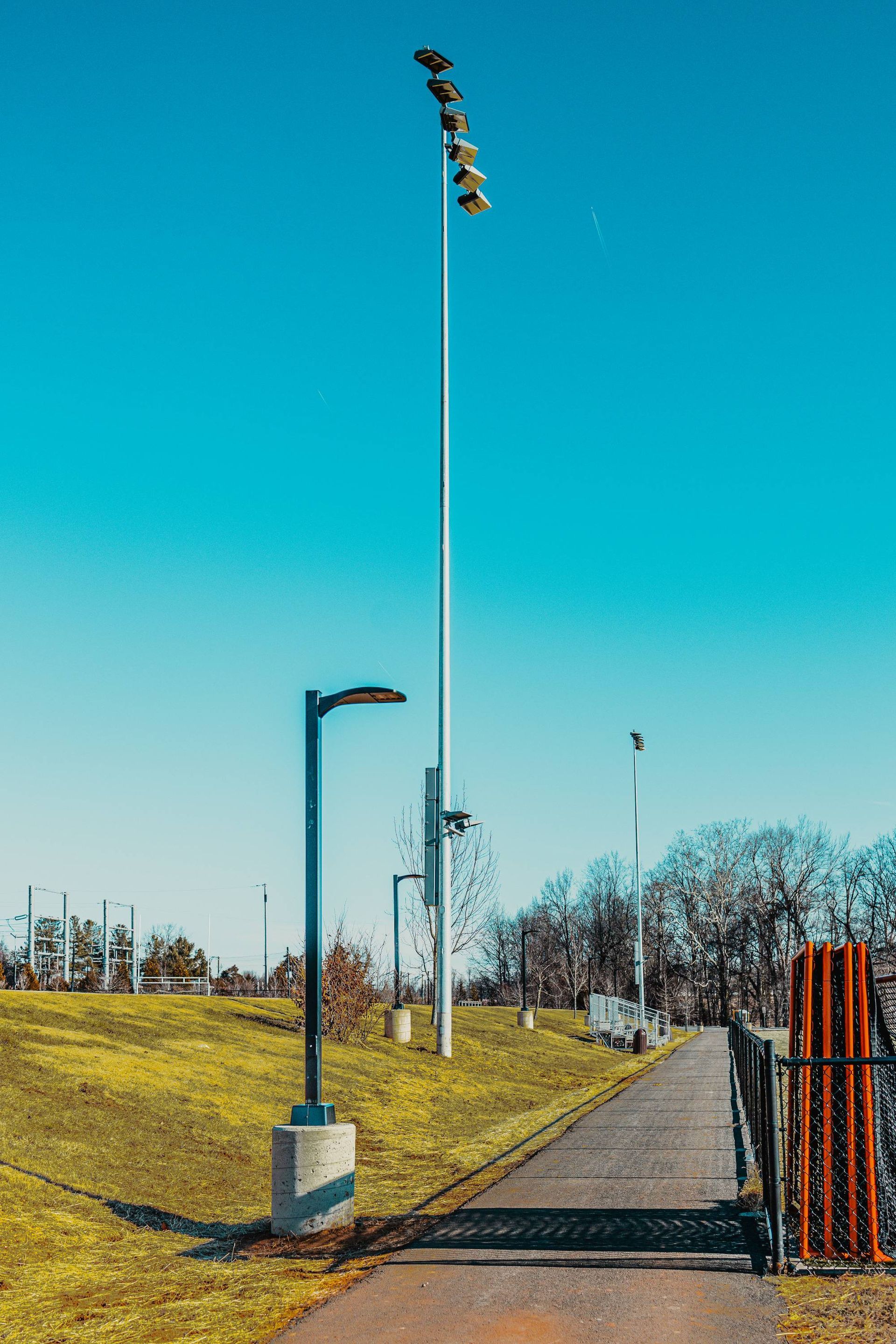 Pathway alongside a grassy hill with tall light poles under a bright blue sky.