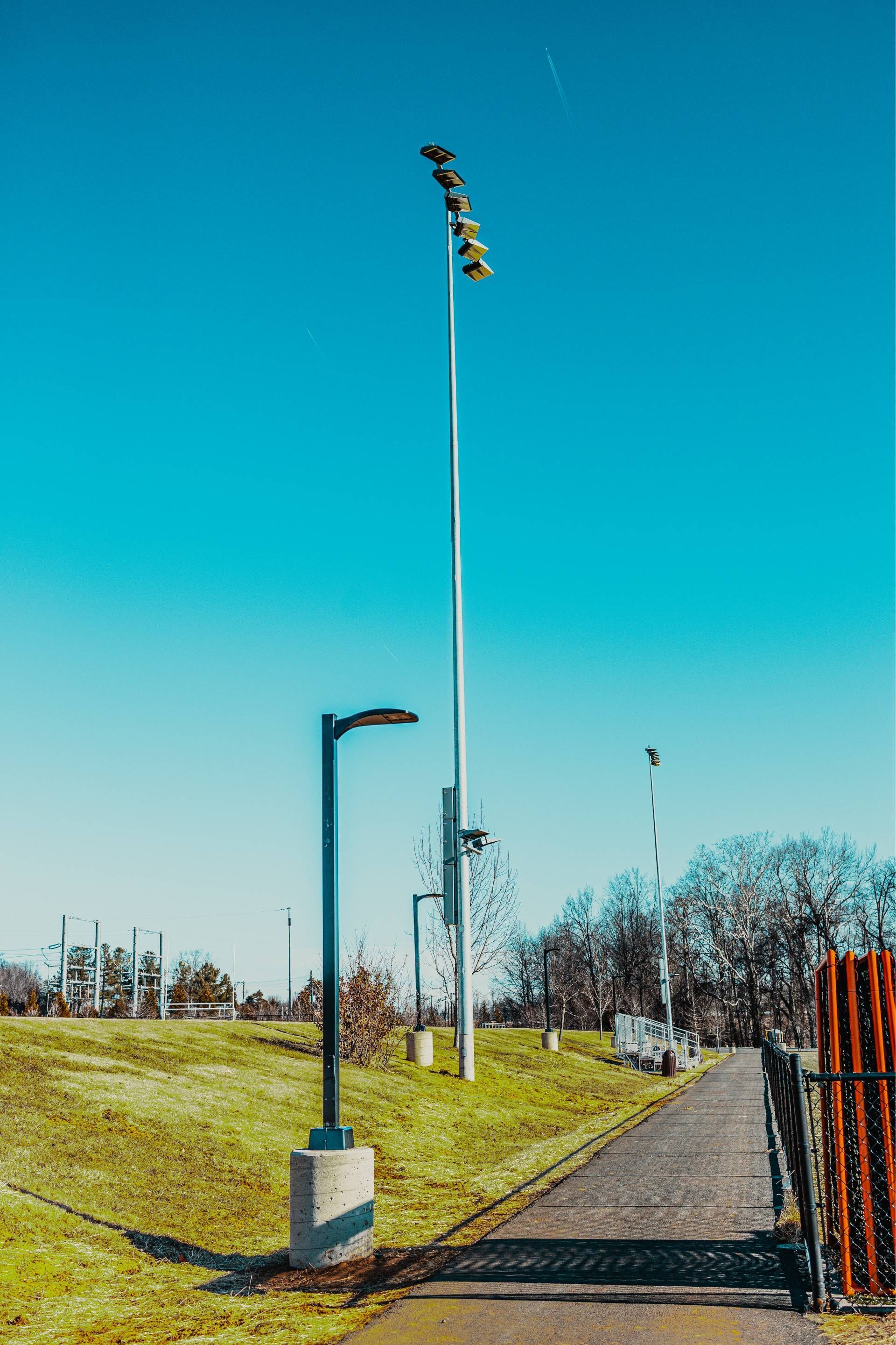 A tall stadium light pole with multiple lights, and a shorter street light on a sunny day.