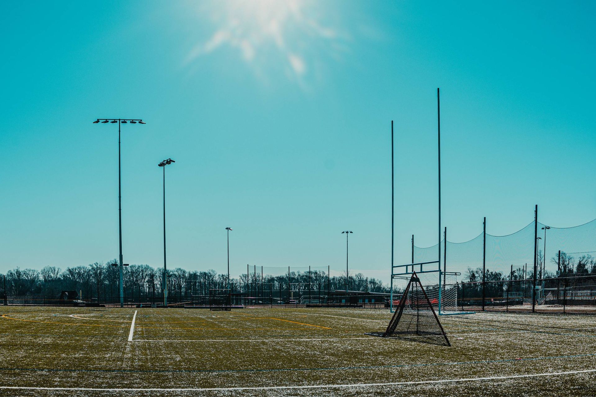 Green sports field under a bright blue sky with stadium lights, goal posts, and netting.