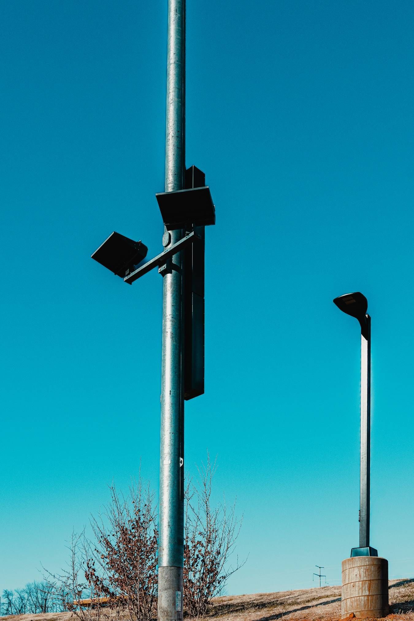 Two tall gray light poles against a clear blue sky. One pole has additional attached equipment.