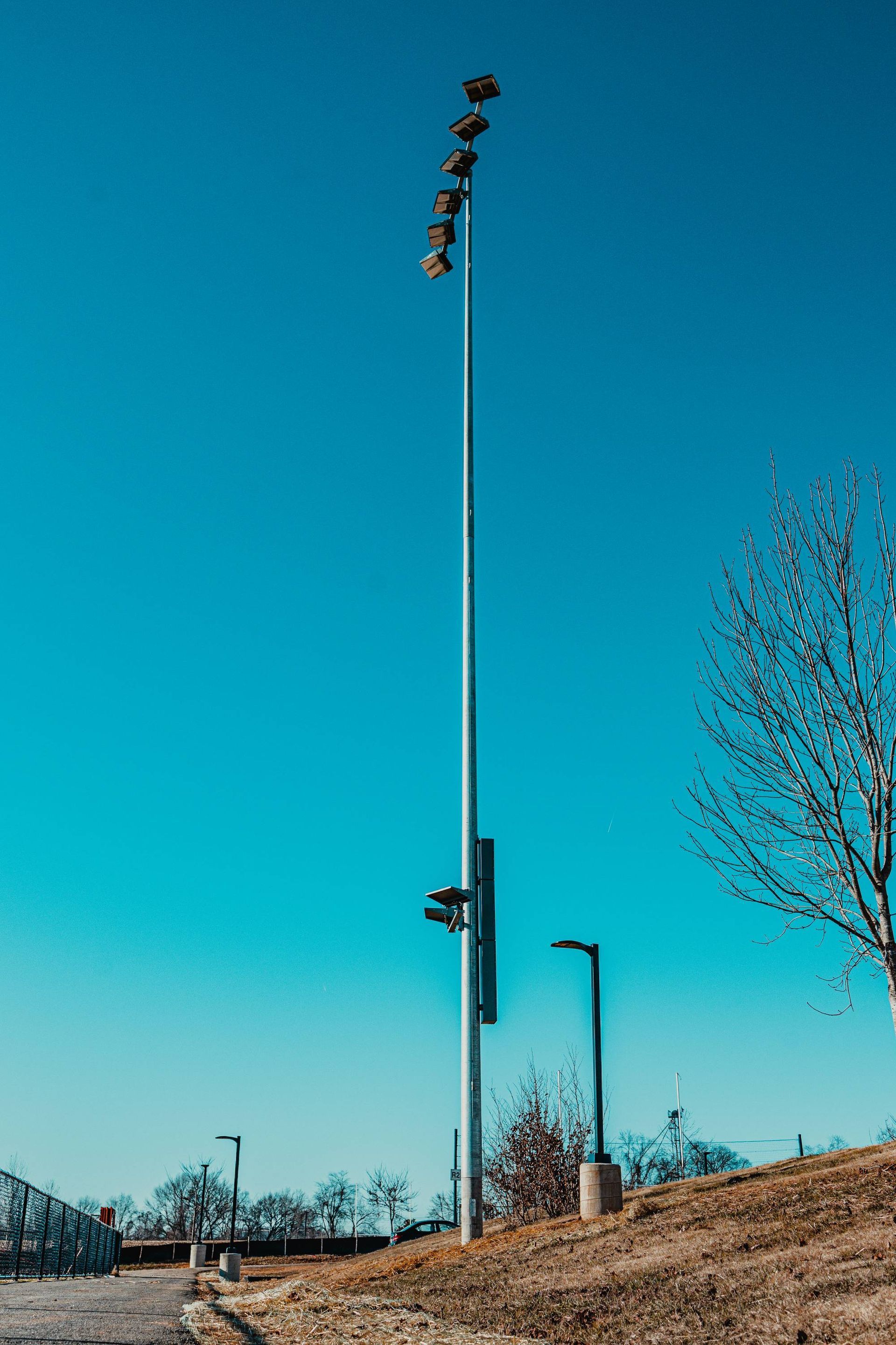Tall metal light pole with several lights against a blue sky. Smaller lights and trees in the background.