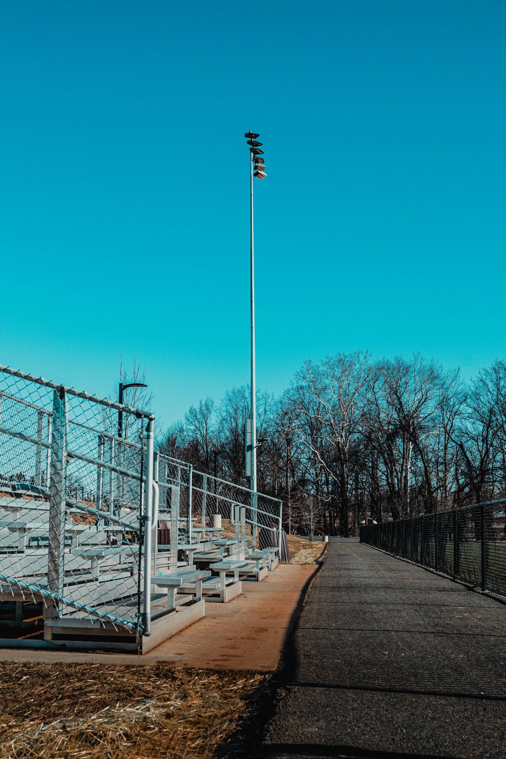 Bleachers next to a chain-link fence and a paved walkway, with a tall light pole against a clear blue sky.