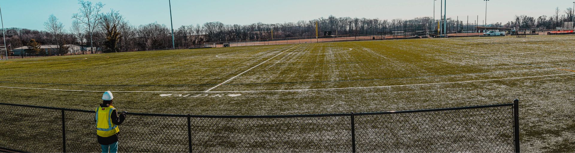 Person in safety vest and hardhat standing near a black fence, with a field and industrial structures in the distance.