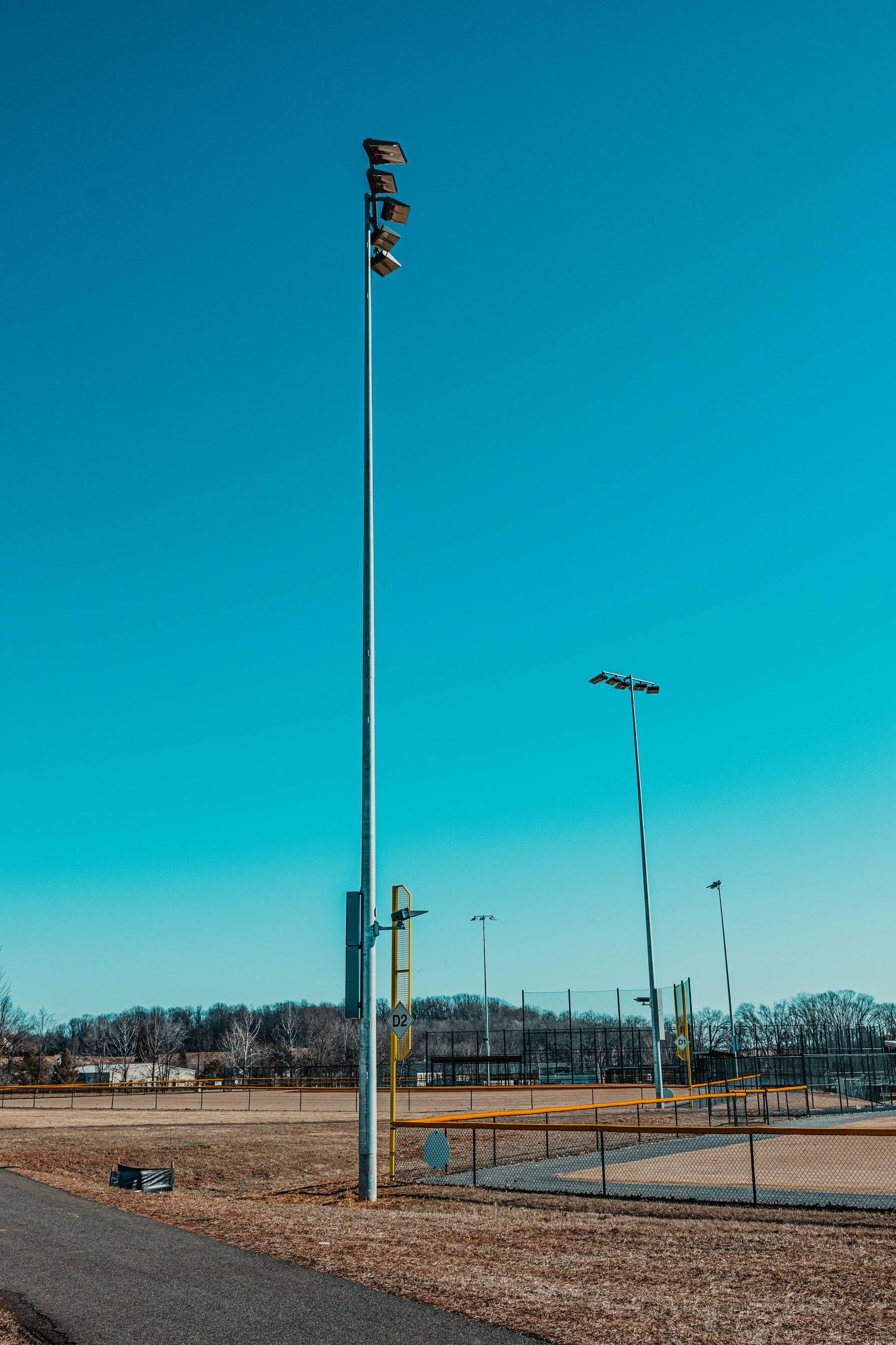 Tall light pole at a baseball field on a sunny day. Blue sky, brown grass, and a path are also visible.