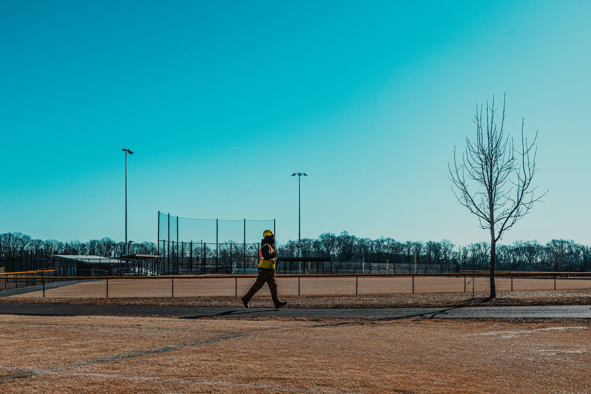 Person walking on a path in front of a baseball field on a sunny day.