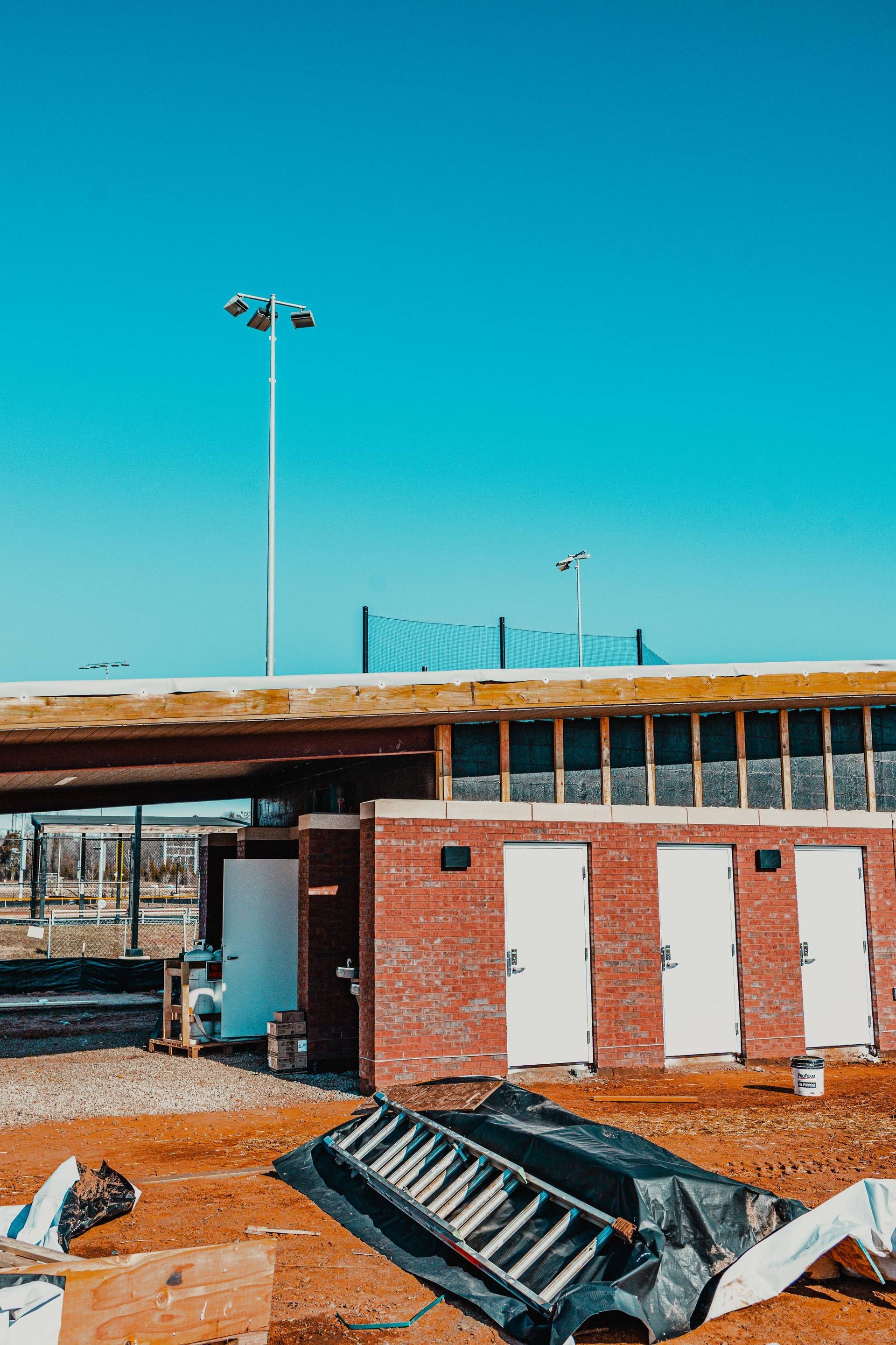 Red brick building with white doors, construction site under clear blue sky, tall light poles.
