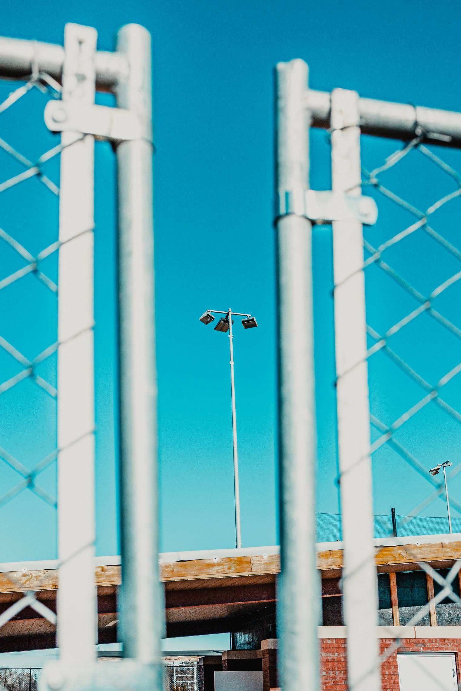 A tall stadium light pole viewed through a chain-link fence, against a blue sky.