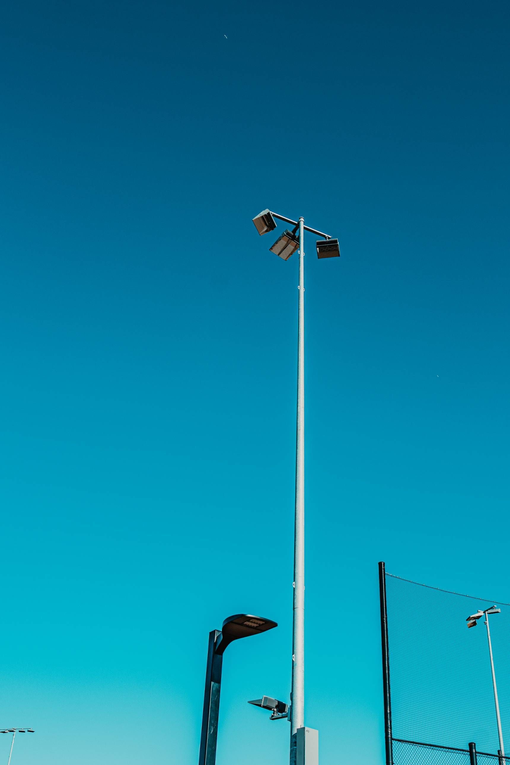 Light poles against a clear, blue sky next to a chain-link fence.