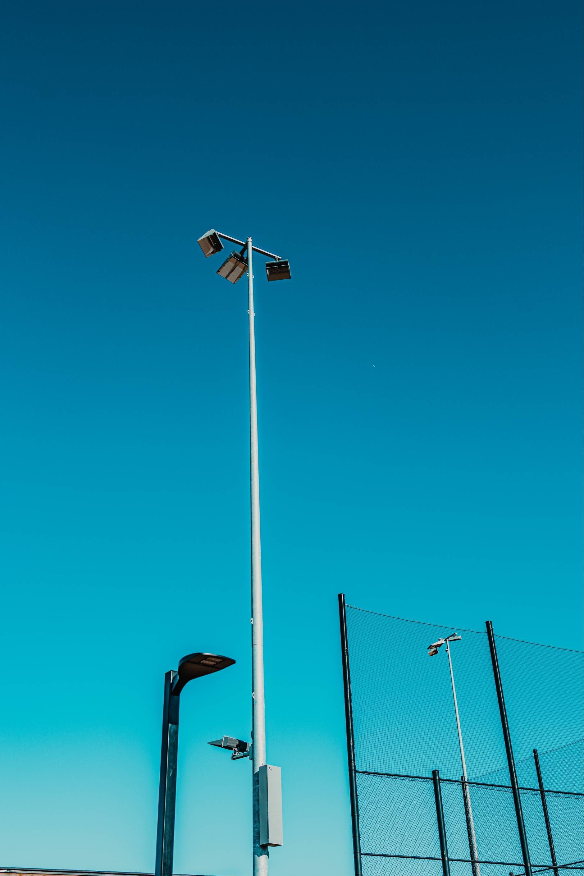 Blue sky with a tall stadium light pole, a smaller light pole, and a chain-link fence.
