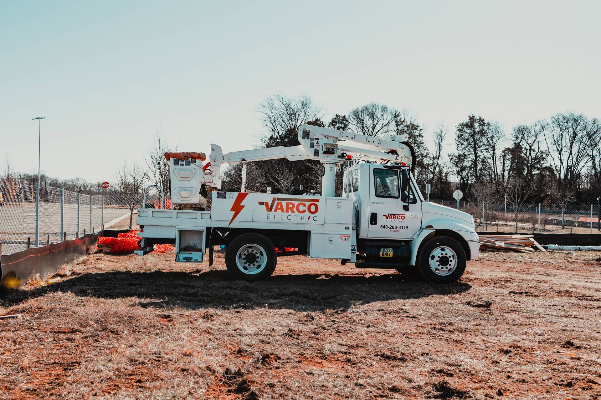 White Varco Electric truck with lift arm parked on dirt in front of a fence.