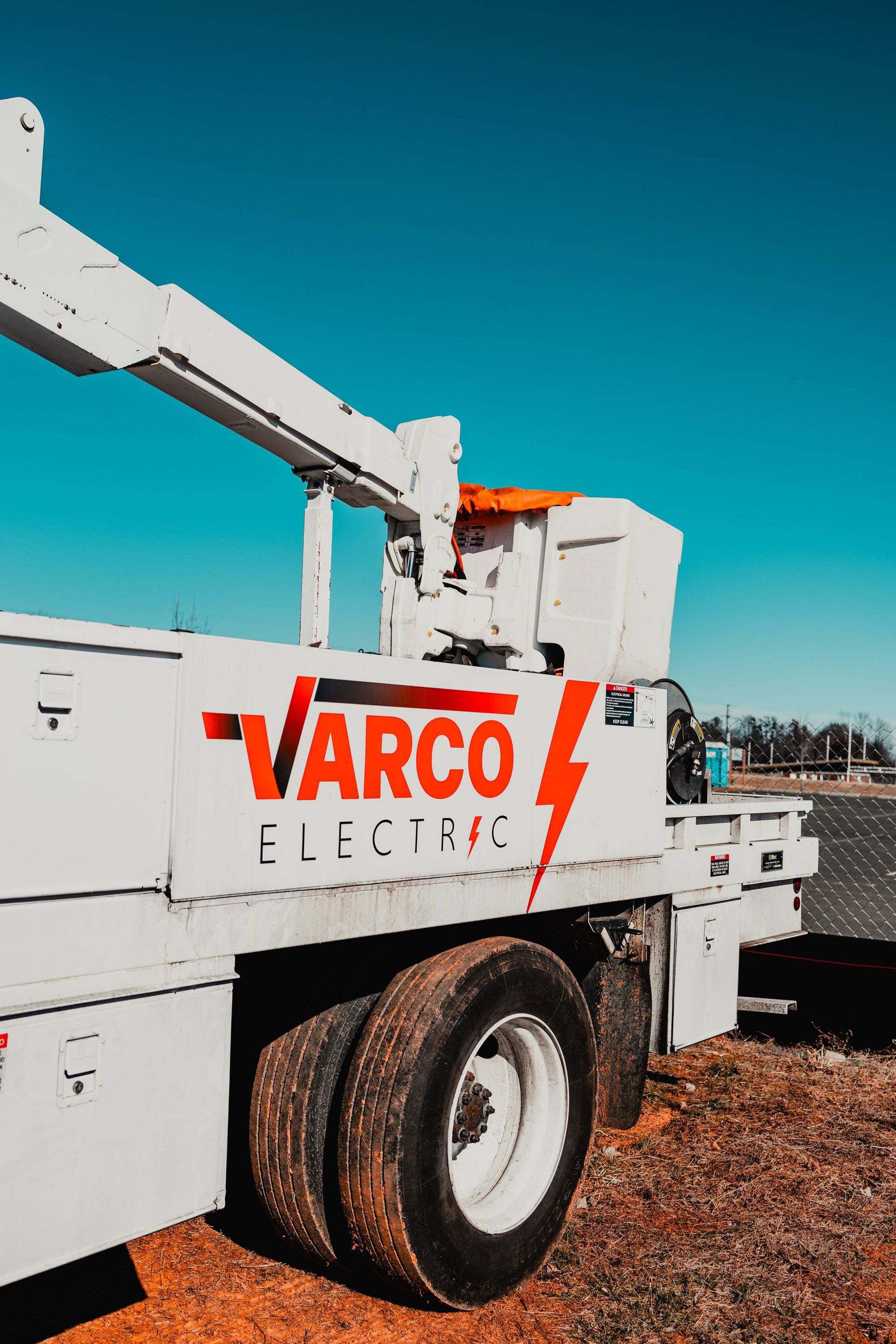 White Varco Electric truck with a red lightning bolt logo and a raised boom, against a blue sky.