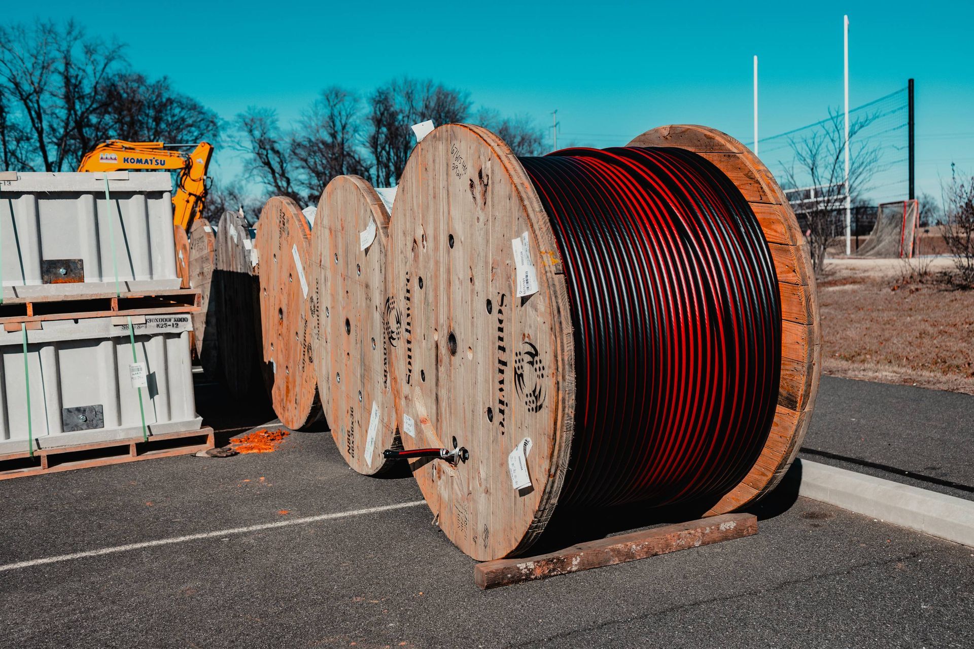 Wooden cable spools sit on asphalt near a building and equipment under a bright blue sky.
