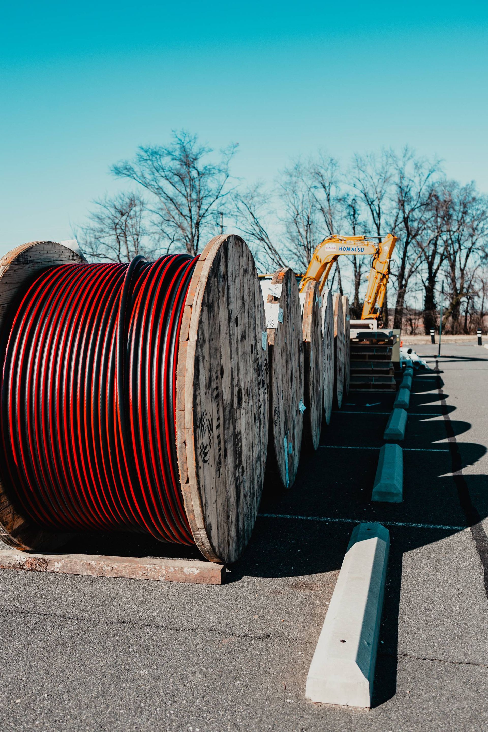 Large spools of red cable on a parking lot, with a yellow excavator in the background under a blue sky.