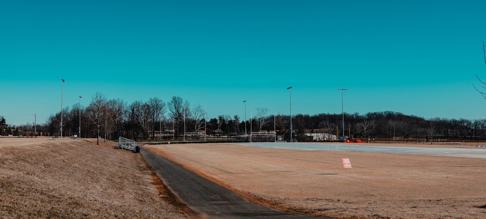An asphalt road leads to a sports field under a blue sky. Bare trees and stadium lights are visible.