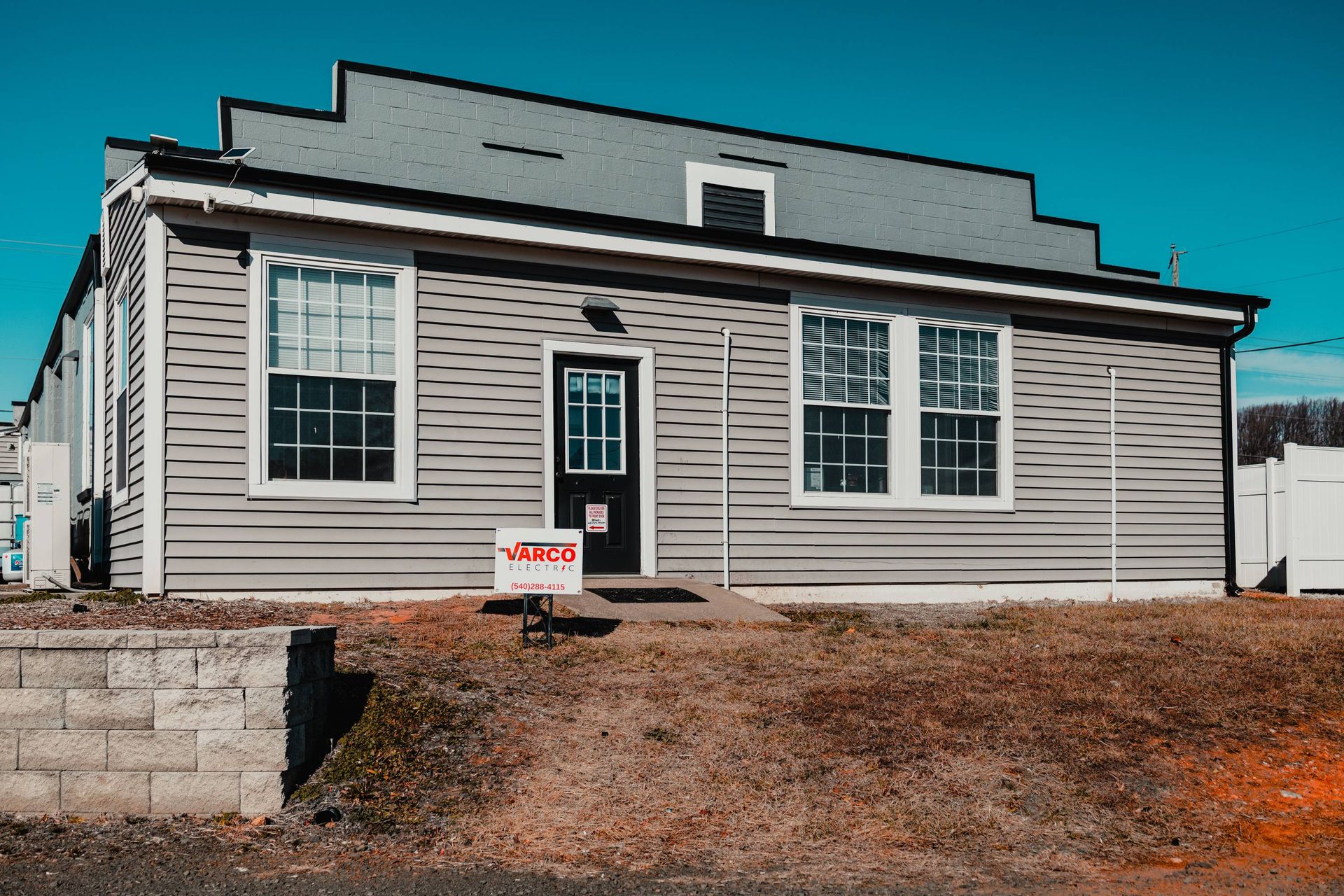 Gray building with white-framed windows and black door under a blue sky. A sign is in front.