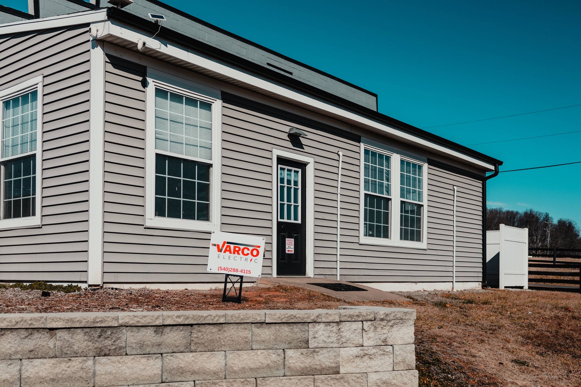 Gray building with white trim and windows, black door, a sign in front, and a blue sky.