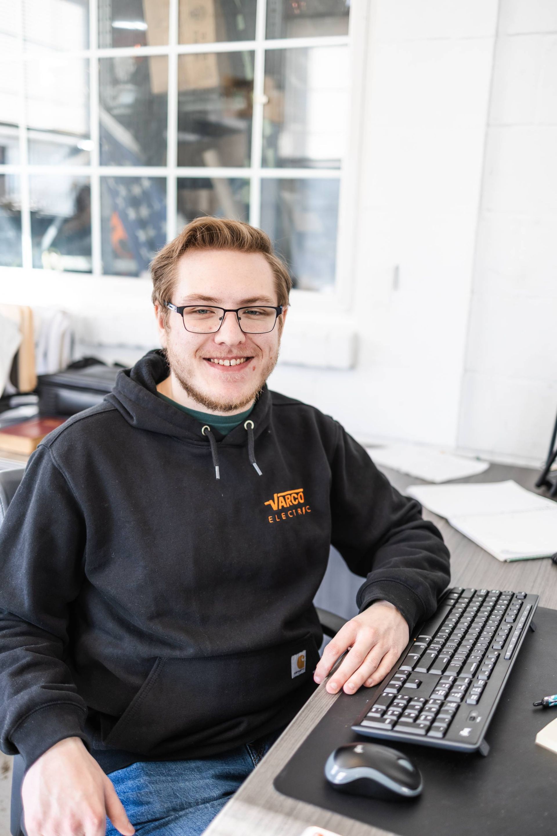 Man in glasses, smiling, wearing a black hoodie, sitting at a desk with a keyboard, near a window.