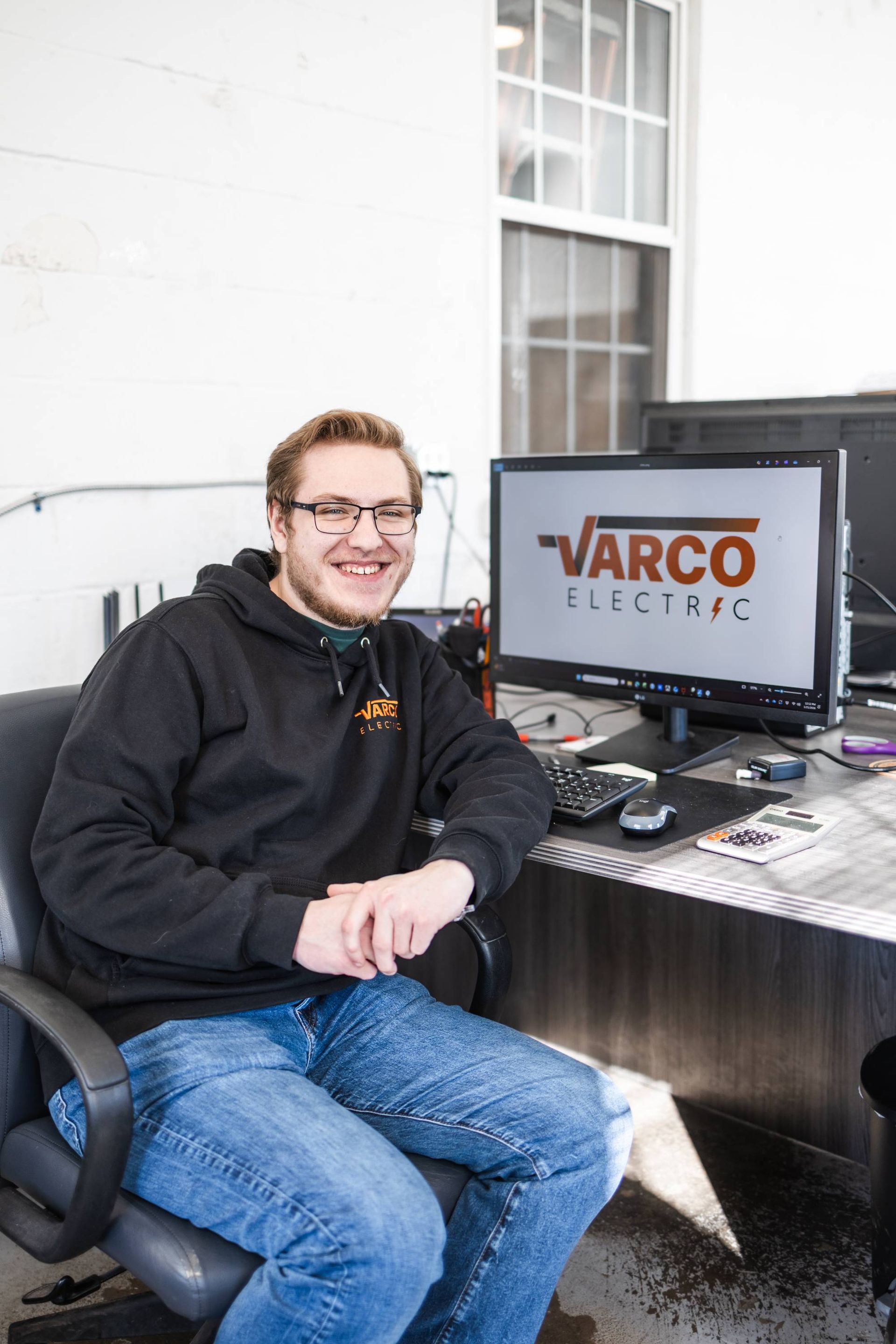 Man sitting at a desk with computer, wearing glasses and a VARCO Electric hoodie, smiling.