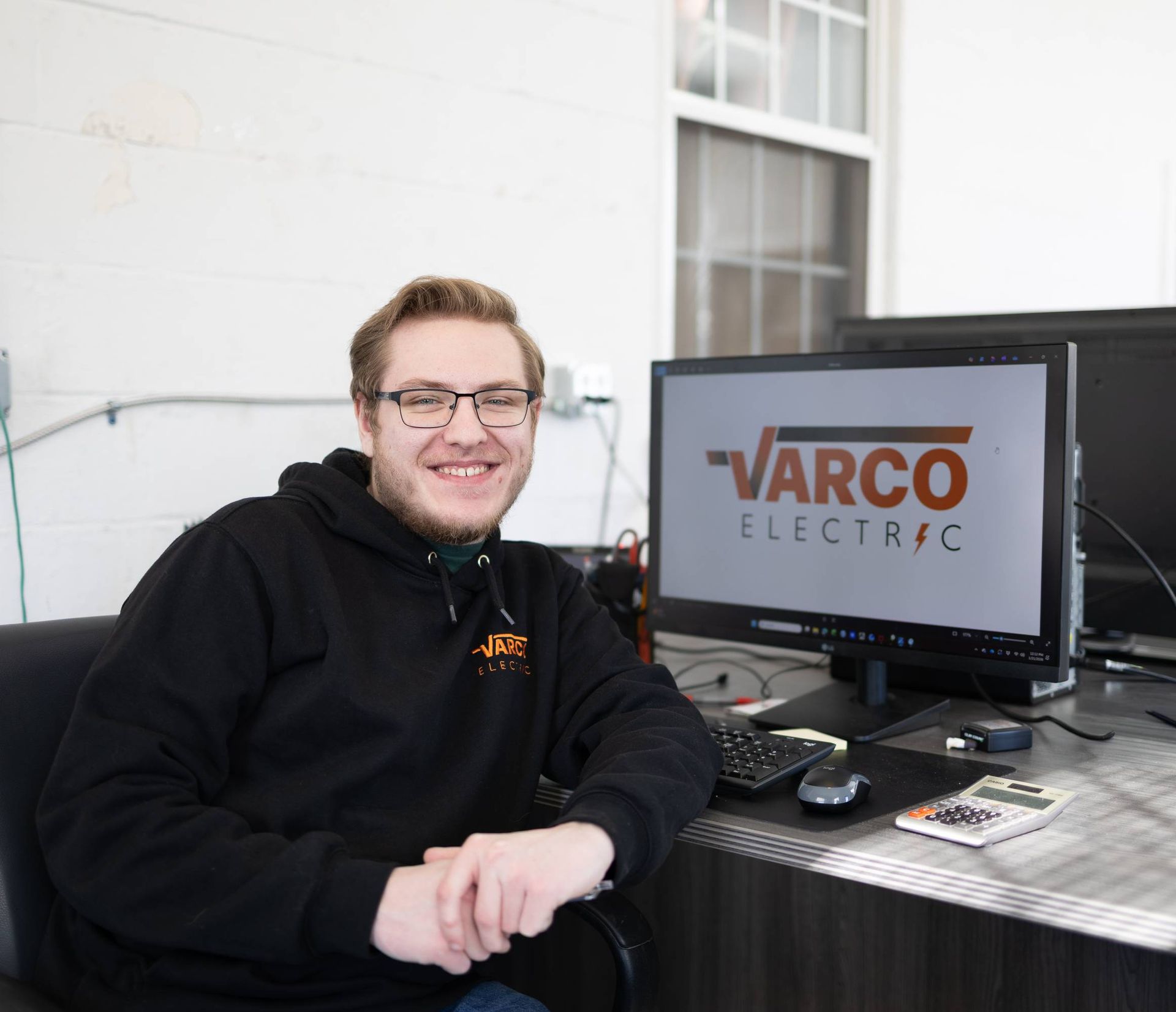 Man in black hoodie, smiling, at desk with computer displaying 