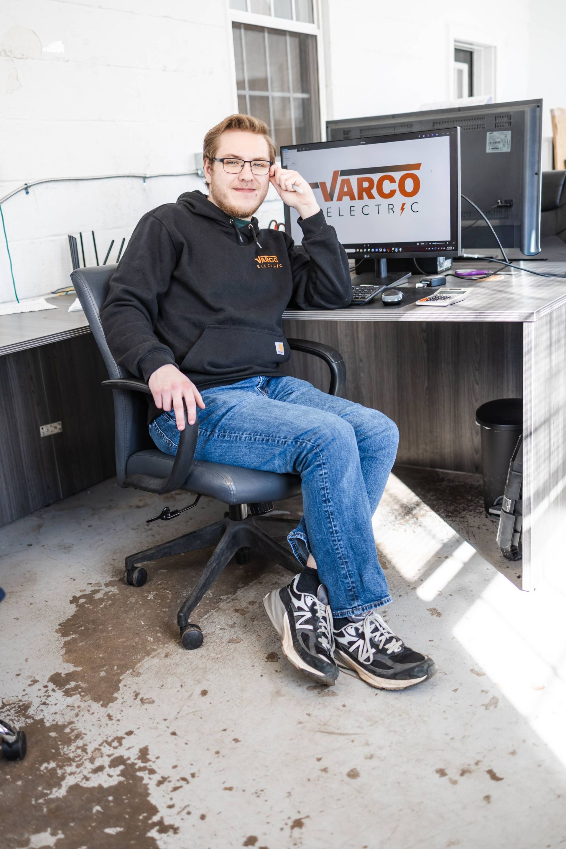 Man in glasses and hoodie sits at a metal desk with computer monitors, outdoors. Water stains on the ground.