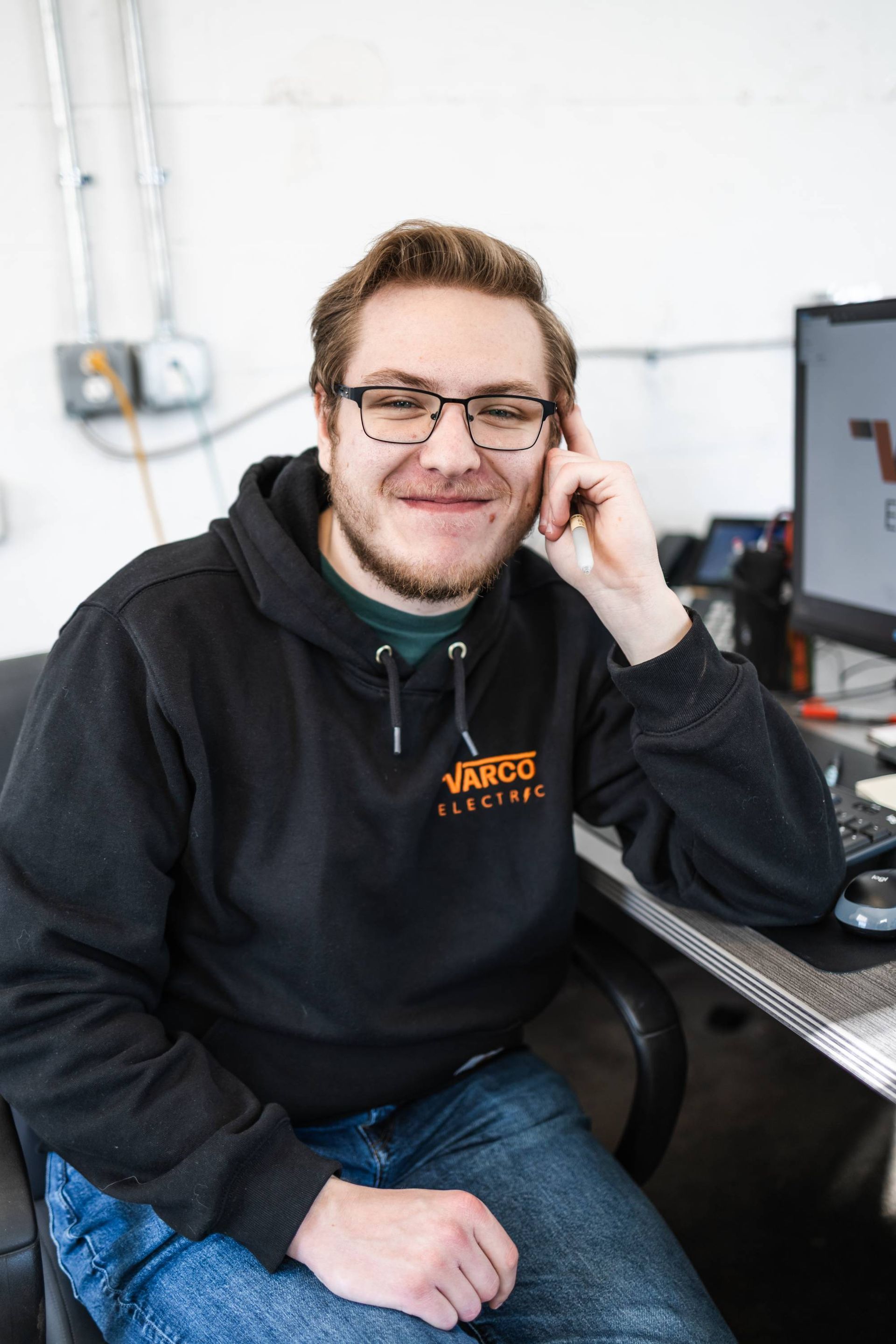 Man in black hoodie smiles, leaning on desk with computer in an office setting.