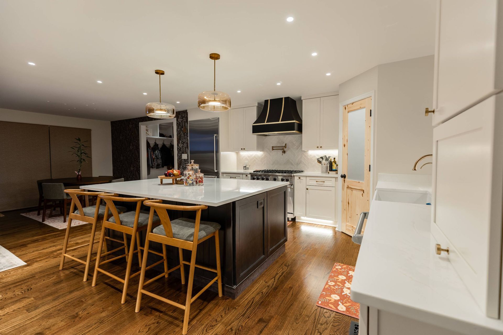 Modern kitchen with island, seating, and overhead lights. Features white cabinets, dark island, and wood floors.
