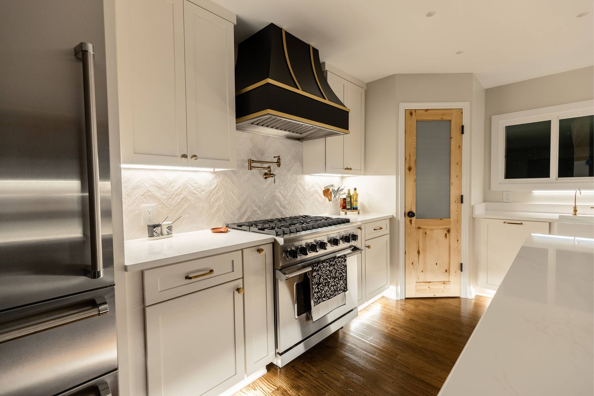 Kitchen with stainless steel appliances, light cabinets, black and gold range hood, and wood floors.