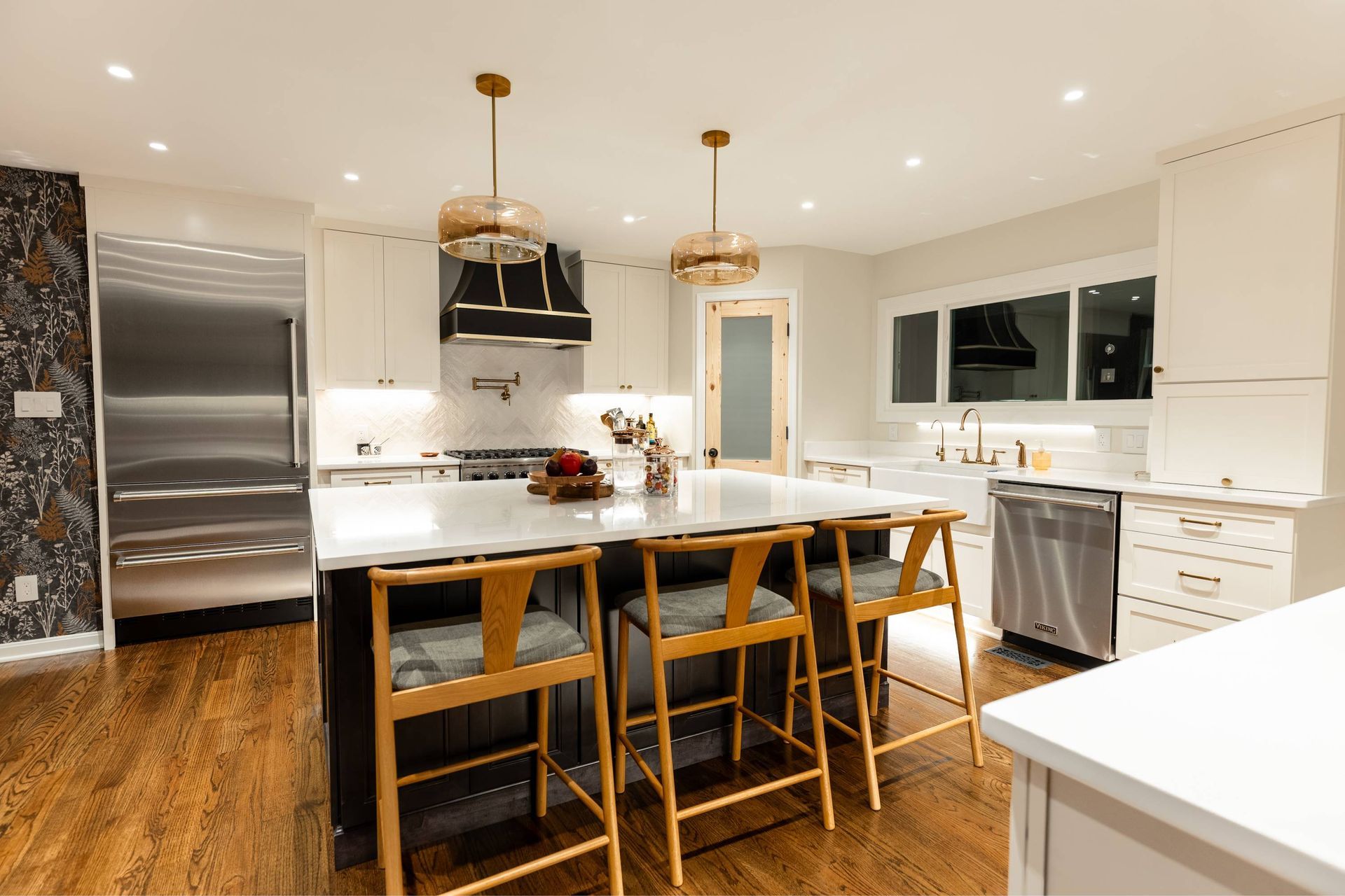 Modern kitchen with island, pendant lights, wooden stools, stainless steel appliances, and dark cabinets.