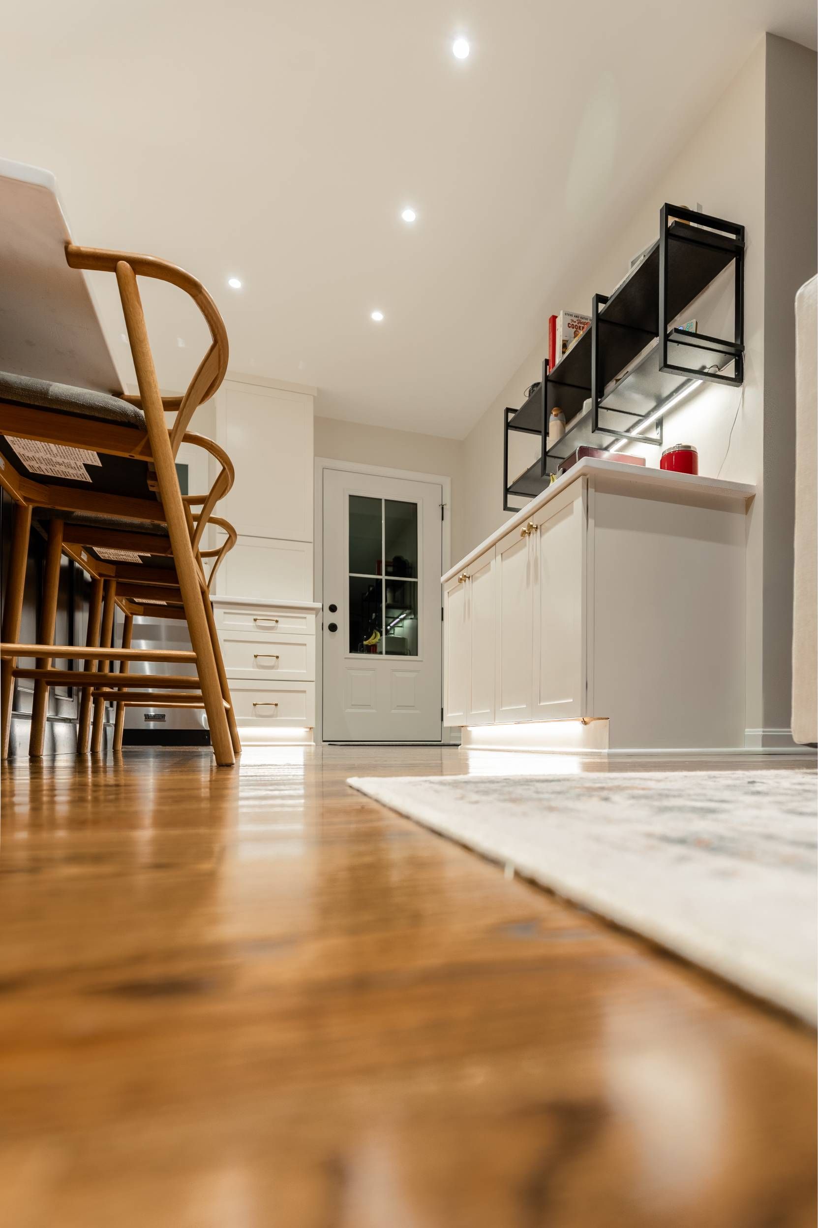 Wooden floor in a bright kitchen with white cabinets, shelves, and a door; chairs along a dining table.