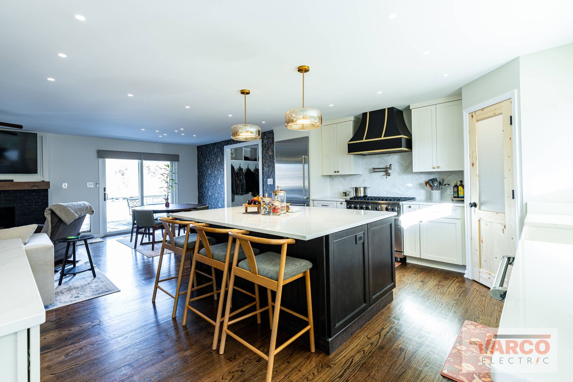 Modern kitchen with dark island and light countertops, stools, white cabinetry, range hood, and pendant lights.