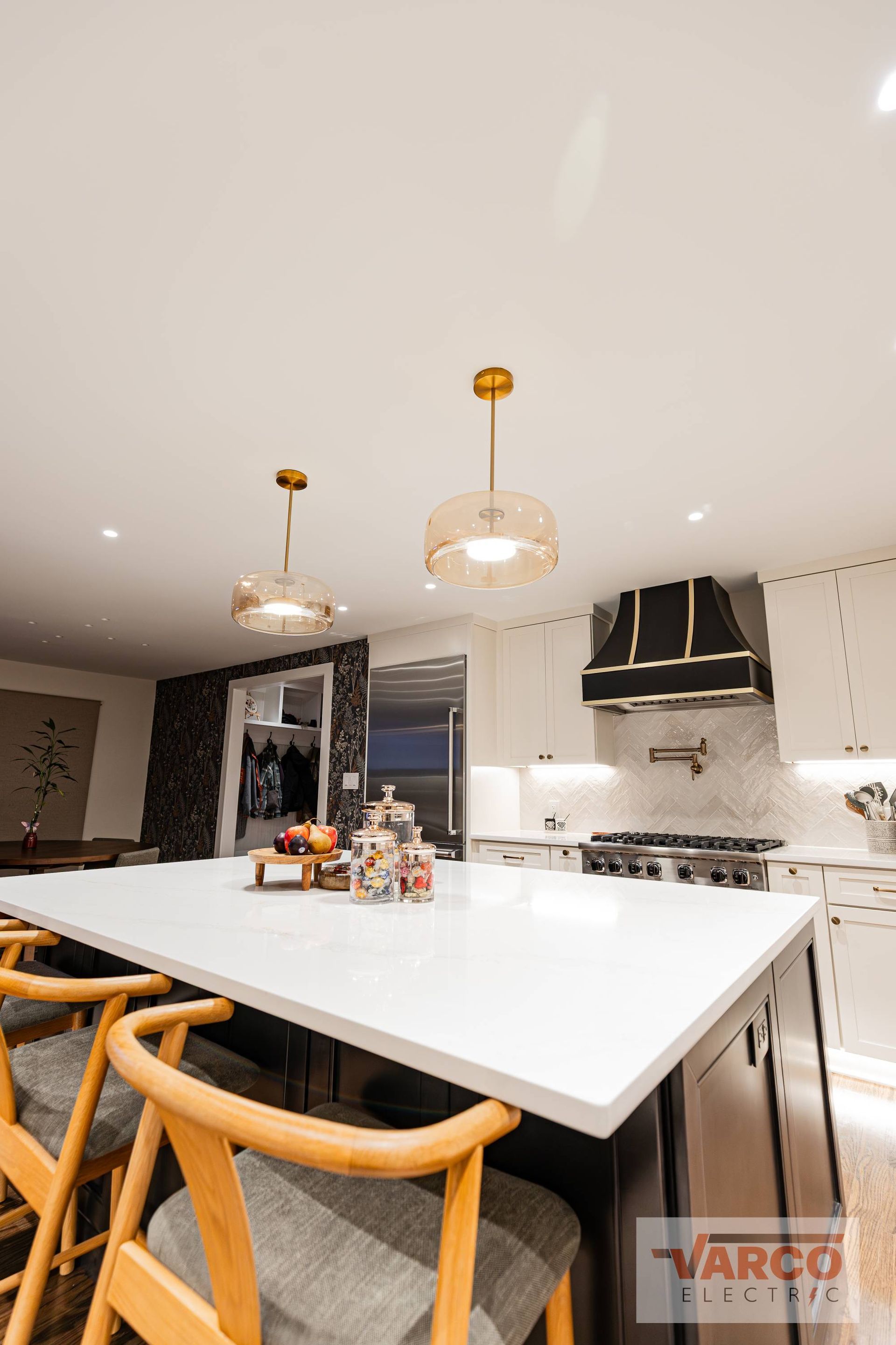 Modern kitchen with island, pendant lights, and black range hood. White countertops and wooden stools.