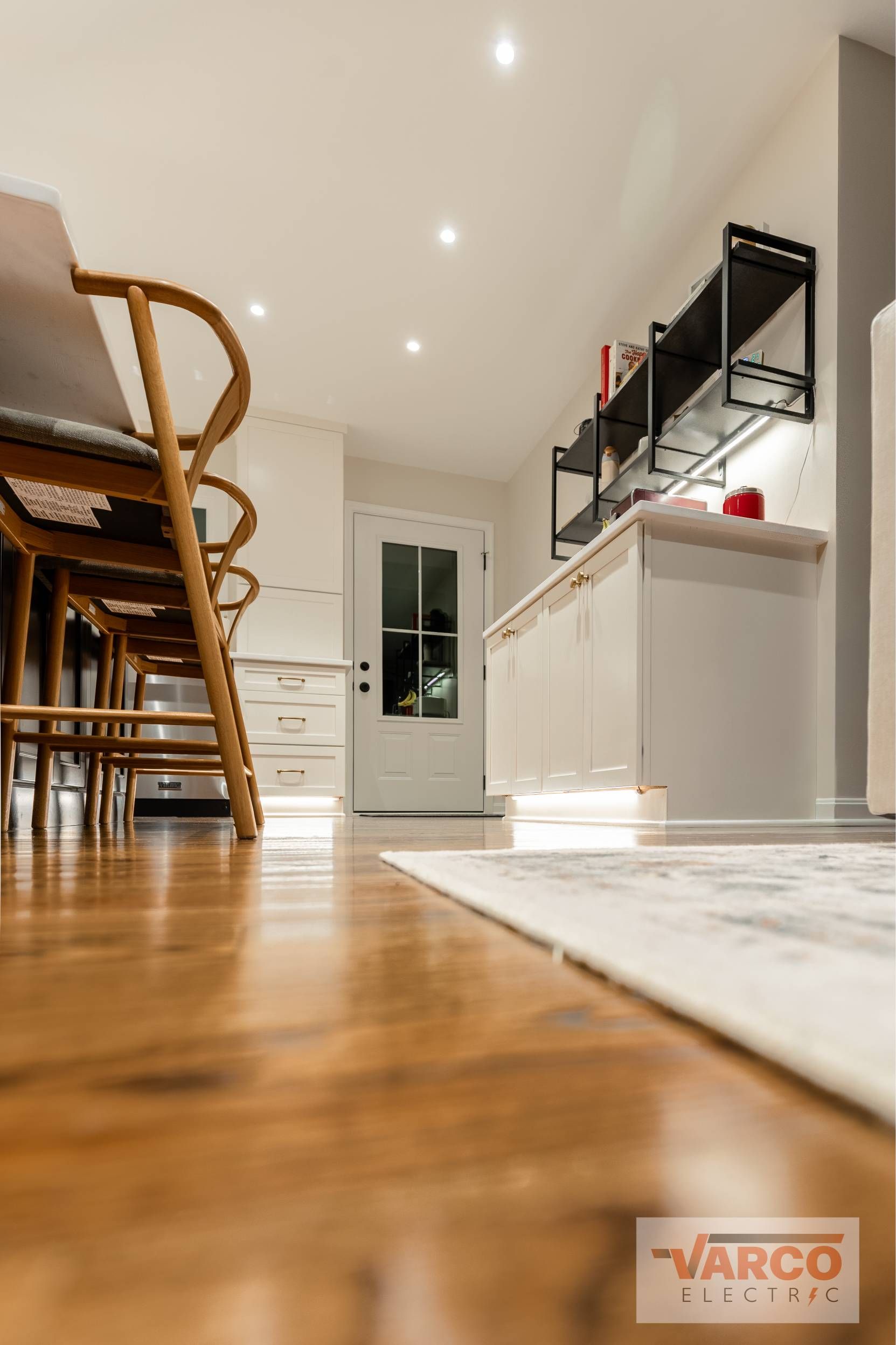 Wooden floor, white kitchen, open shelves, white door, chairs at a table, recessed lighting.