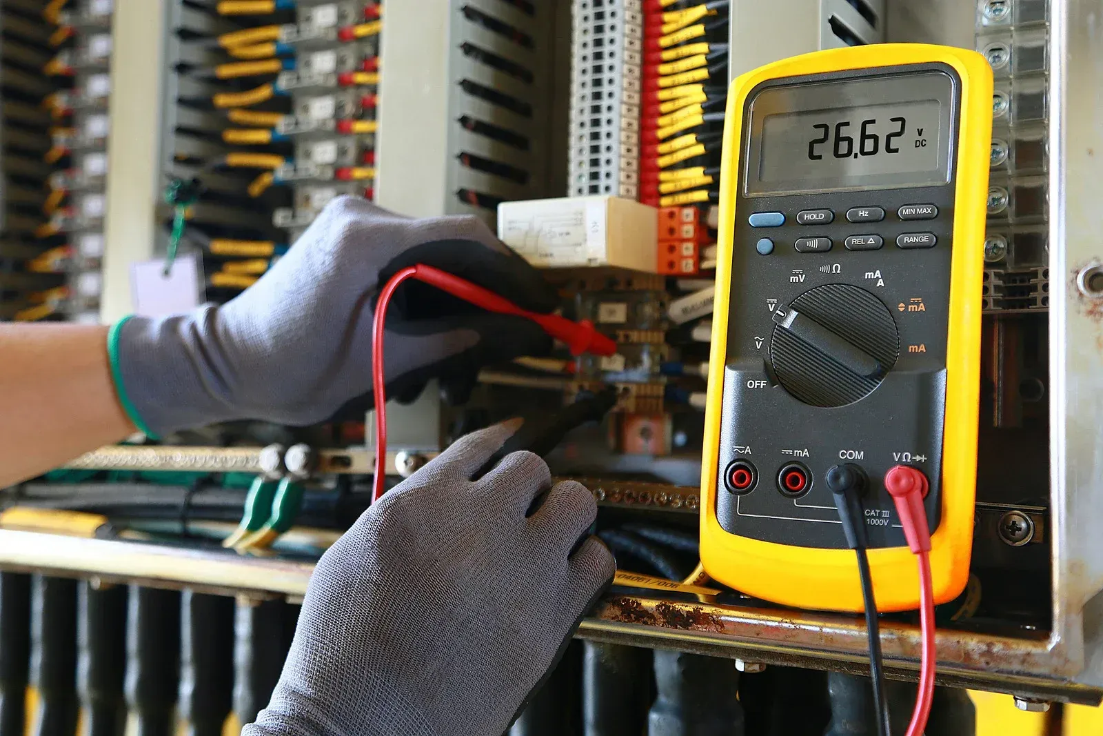 Electrician testing electrical panel with a multimeter; hands in gloves, showing 26.62 volts.