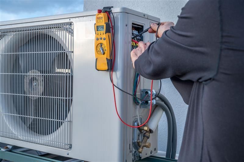 Person in black shirt repairing an AC unit with a multimeter attached. Outdoors, gray AC unit.