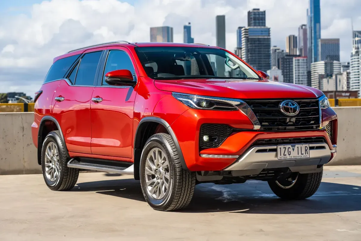 A red toyota fortuner is parked in a parking lot in front of a city skyline.